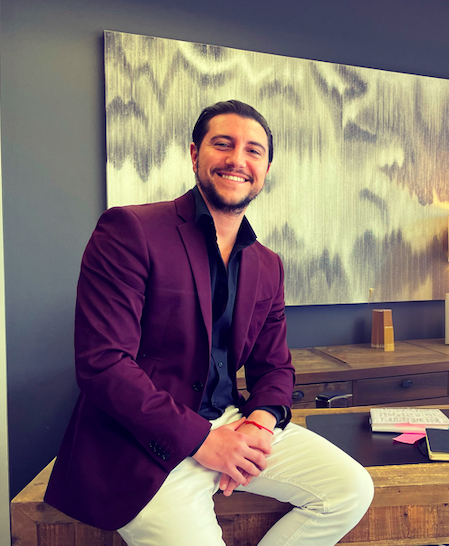 Man with dark hair and beard smiling, sitting on a wooden table, wearing a purple blazer and white pants, in a modern room.