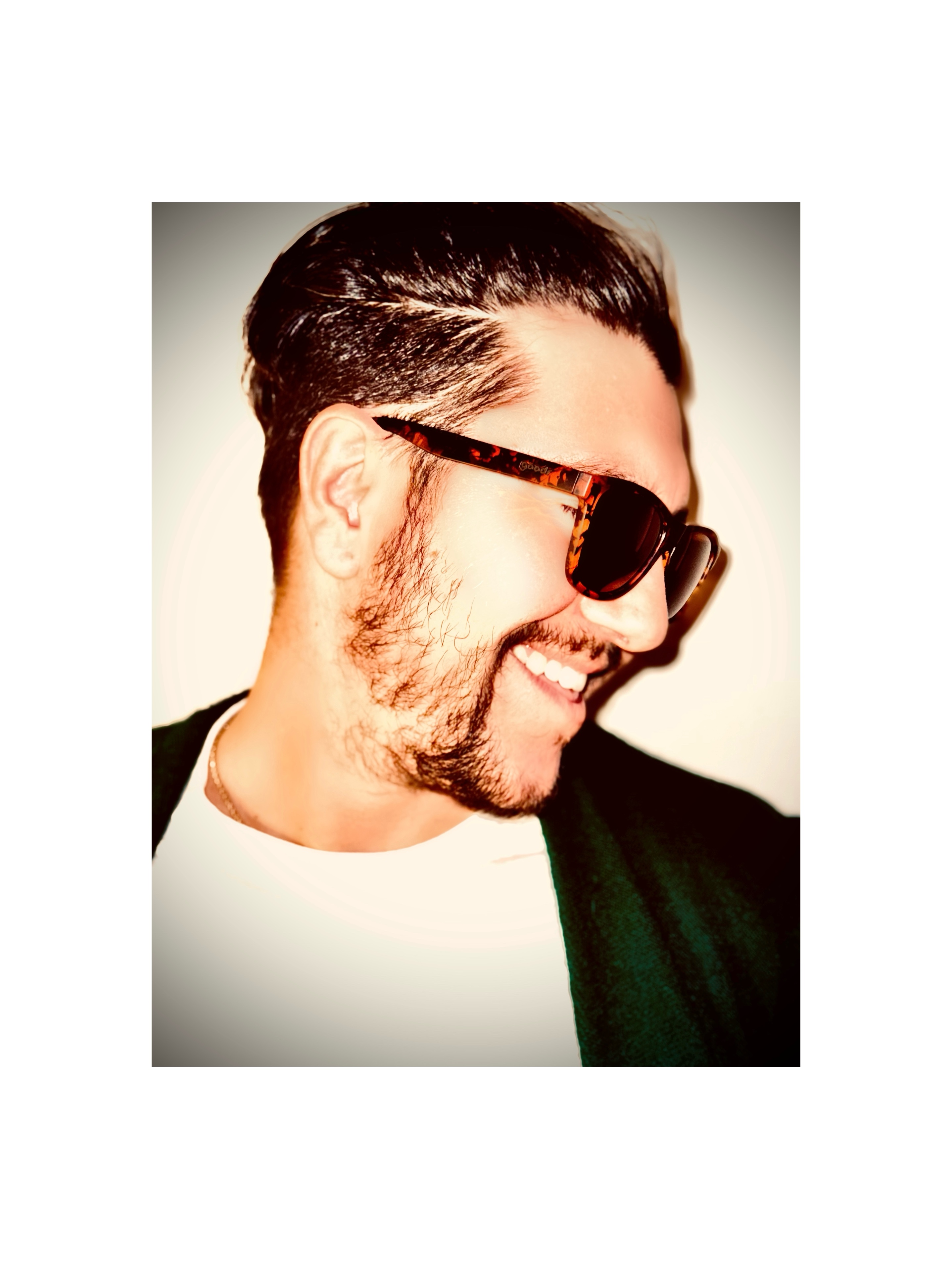 Man with dark hair, beard, sunglasses, smiling, wearing a white shirt and dark jacket, facing right.