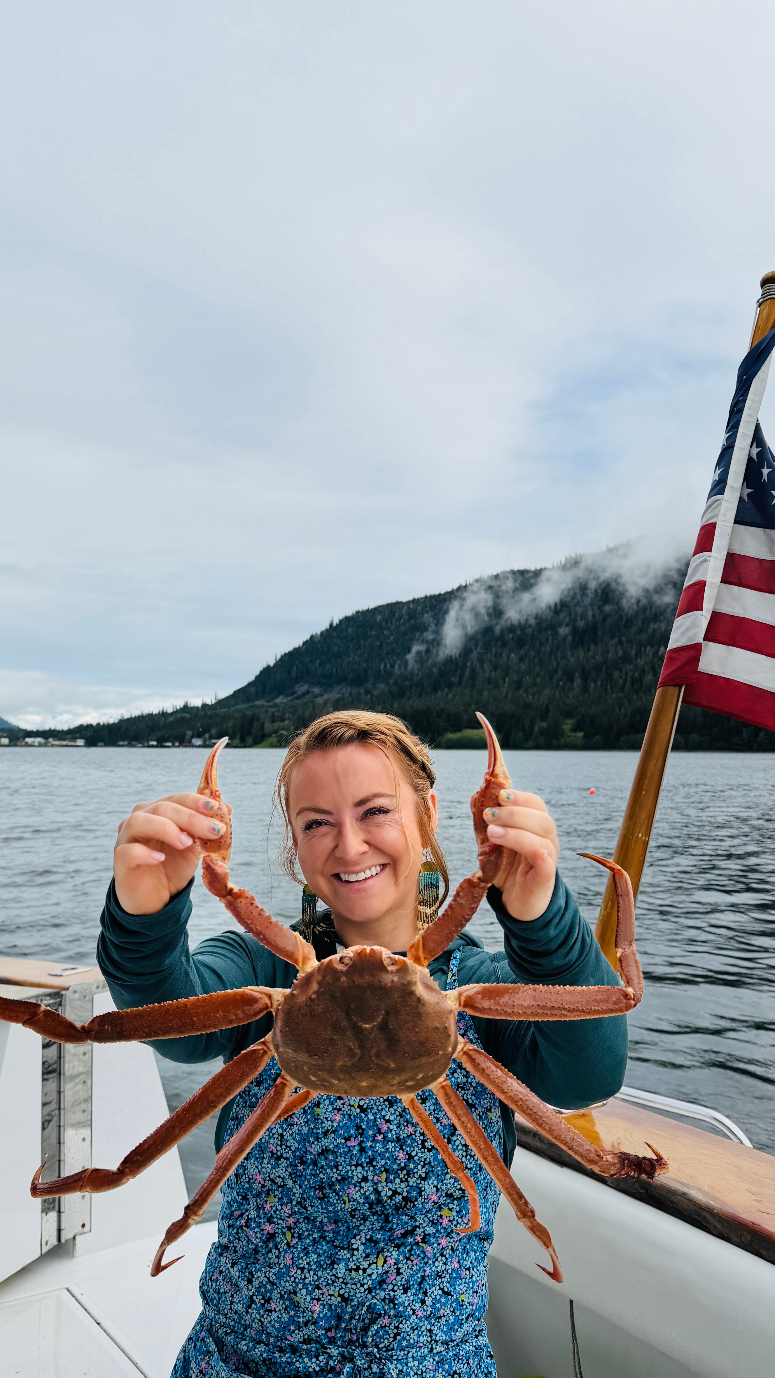 Woman holding a large crab on a boat with water, mountains, and an American flag in the background.