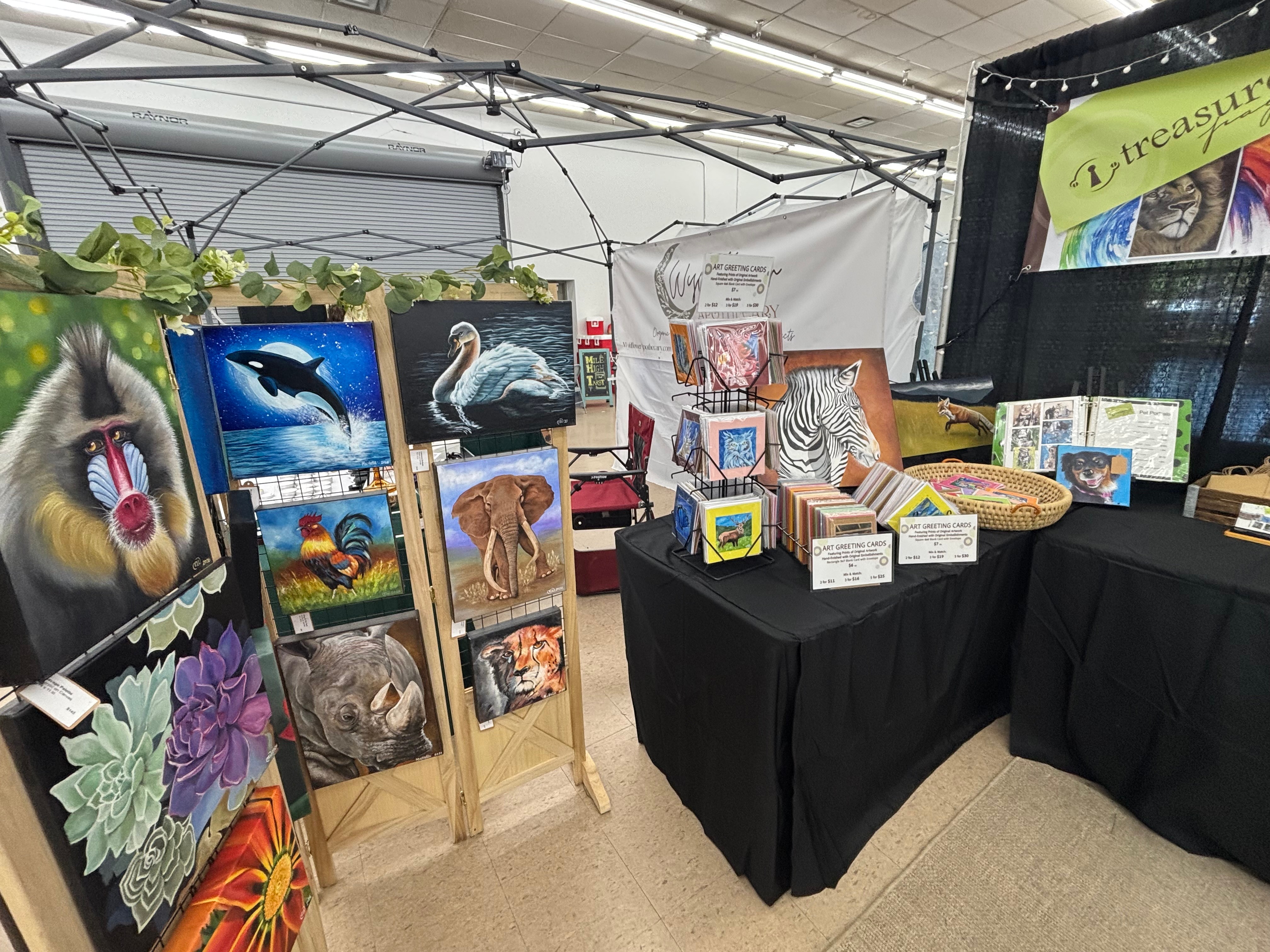 Art display with paintings of animals and nature, tables with art supplies and framed artworks, inside a tent or booth.