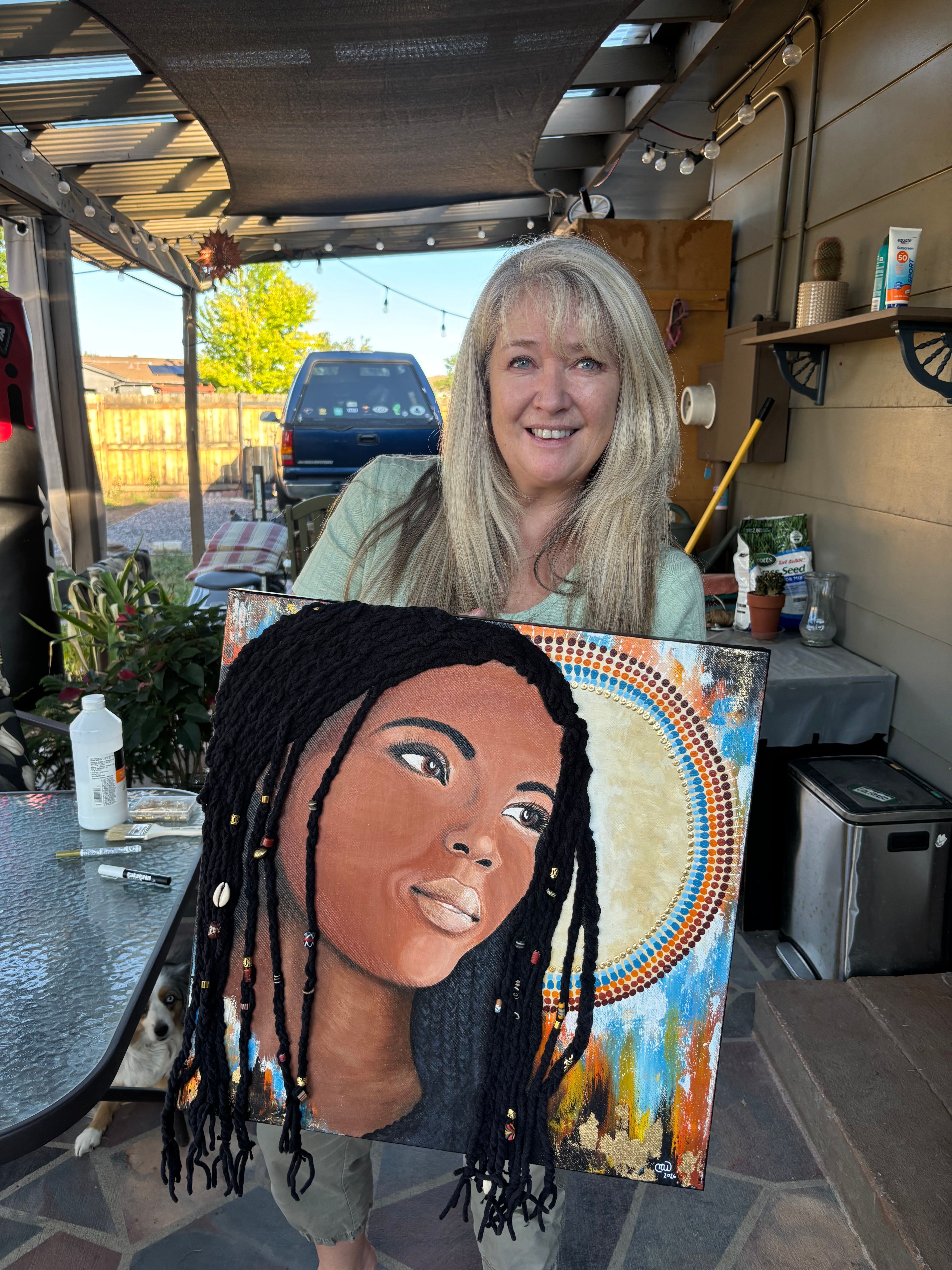 Two women, one with long blonde hair and glasses, and the other with long black dreadlocks, holding a colorful portrait painting.