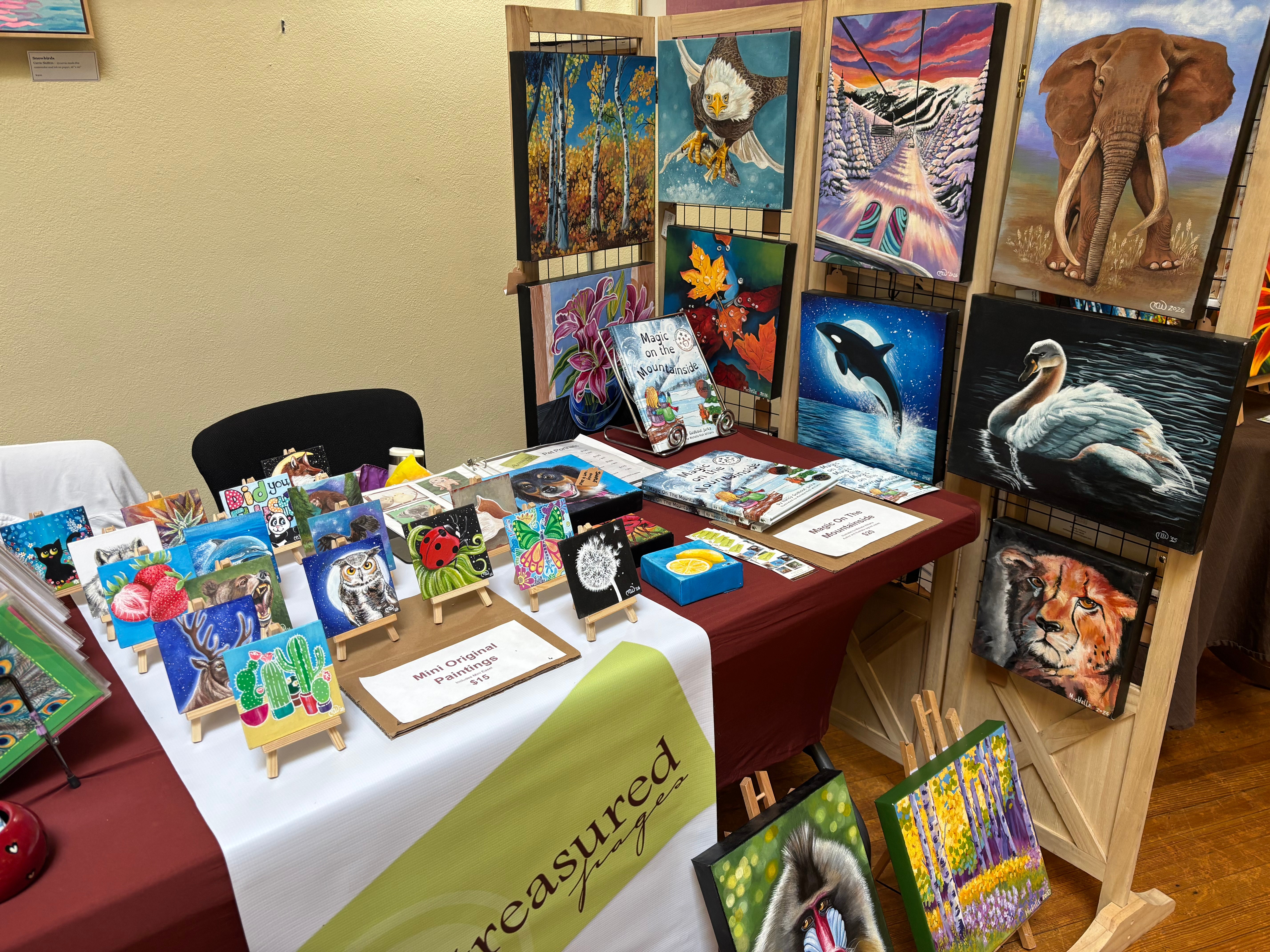 Table with art supplies and paintings, display of colorful artwork, and a wooden stand with animal paintings, including an elephant and a swan.