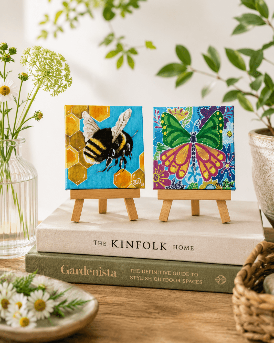 Two small paintings of a bee and a butterfly on mini easels, surrounded by plants and books, on a wooden surface.