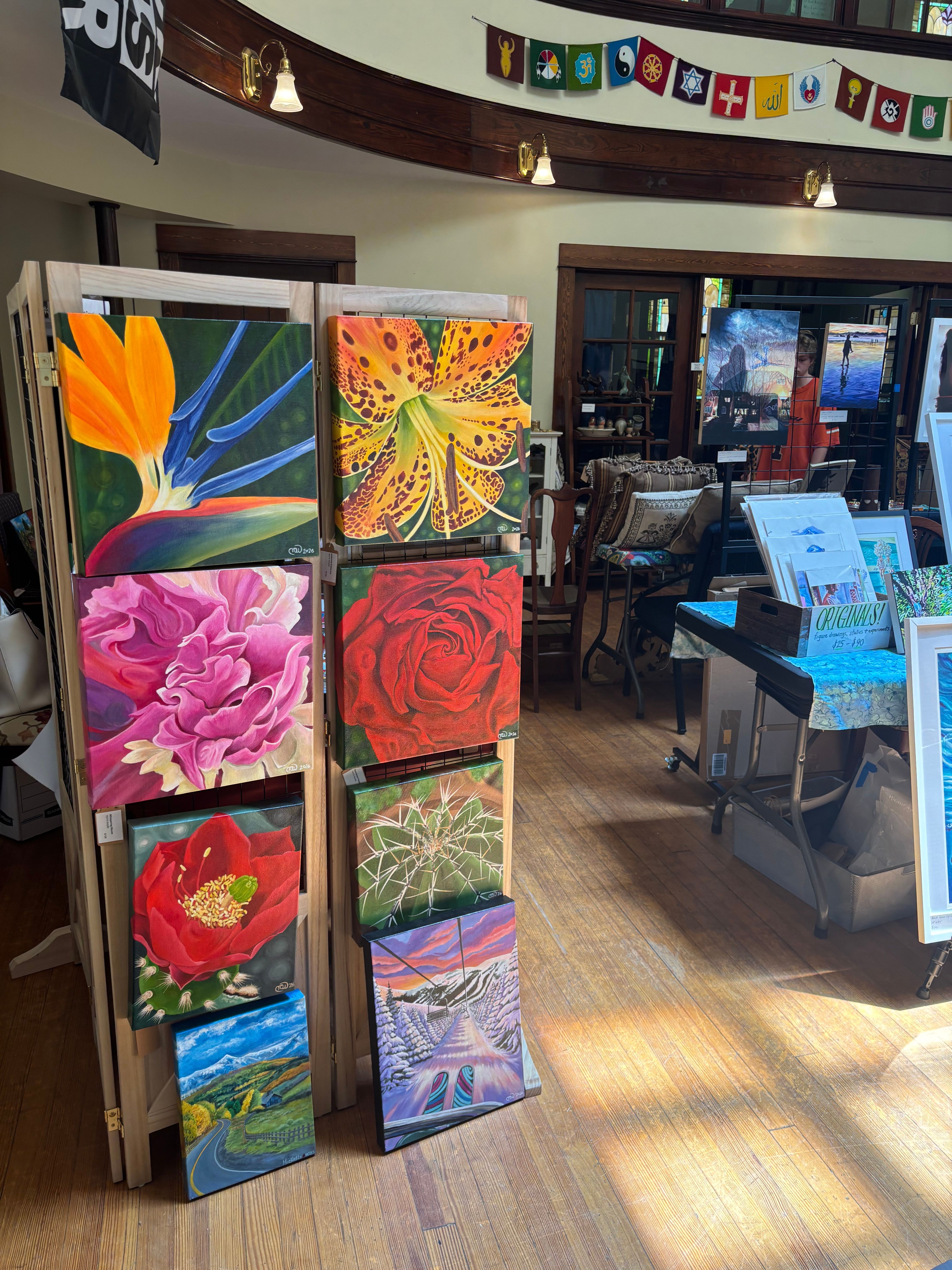 Table with colorful paintings displayed in a room with wooden floor and a window in the background.