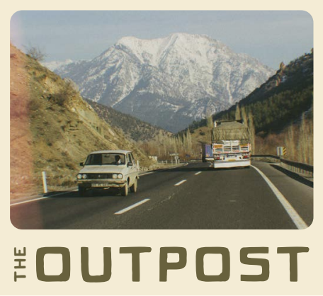 Two vehicles driving on a mountain road with snow-capped peaks in the background, trees on the side, and a clear sky.