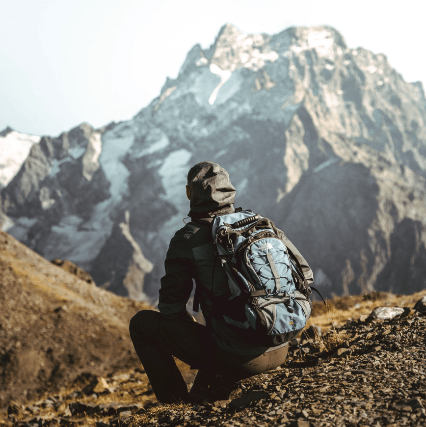 Person with backpack sitting on rocky ground in mountain landscape with snow-capped peaks in background.