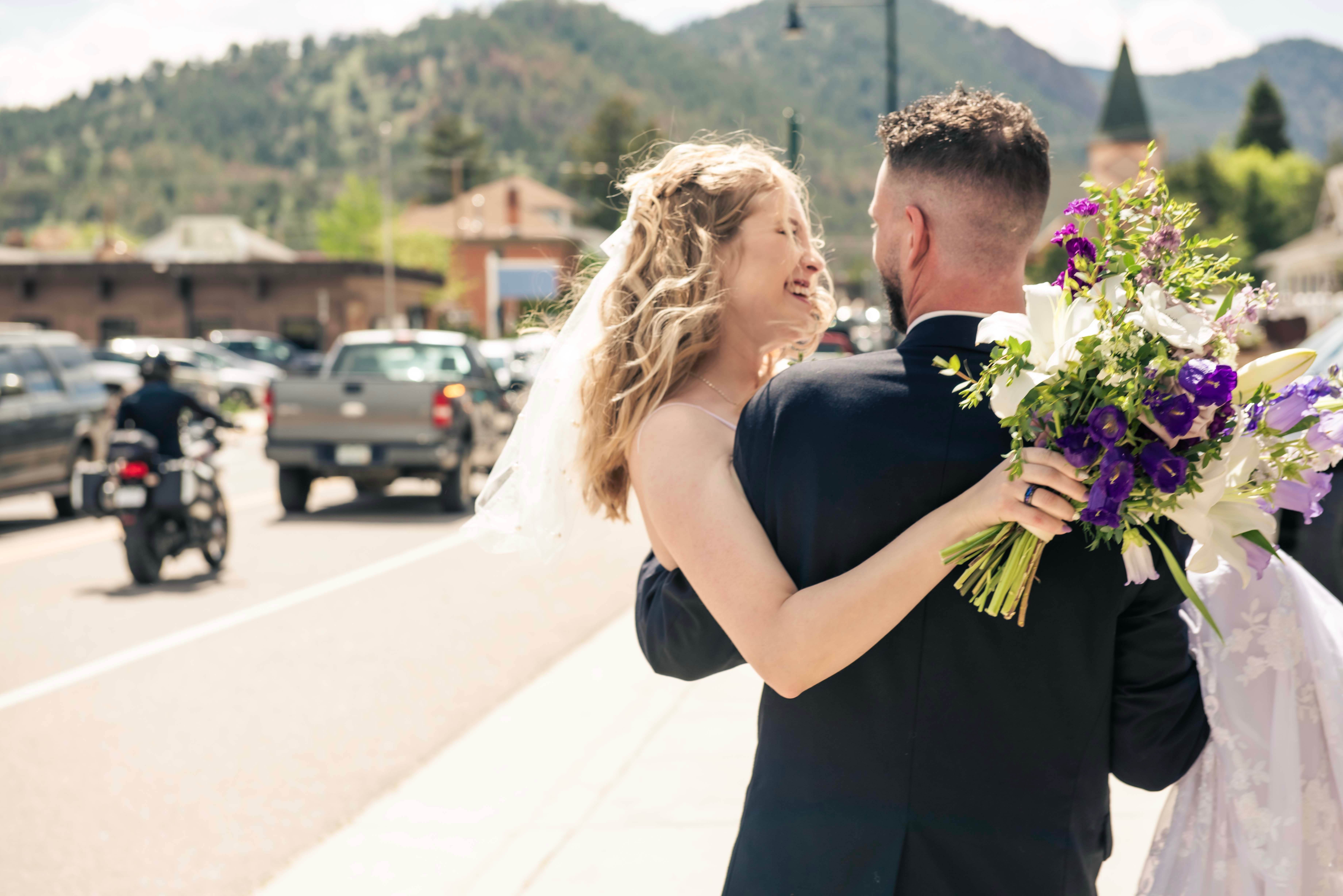 A couple dancing outdoors with a city street, cars, and mountains in the background, the woman holding a bouquet of flowers.