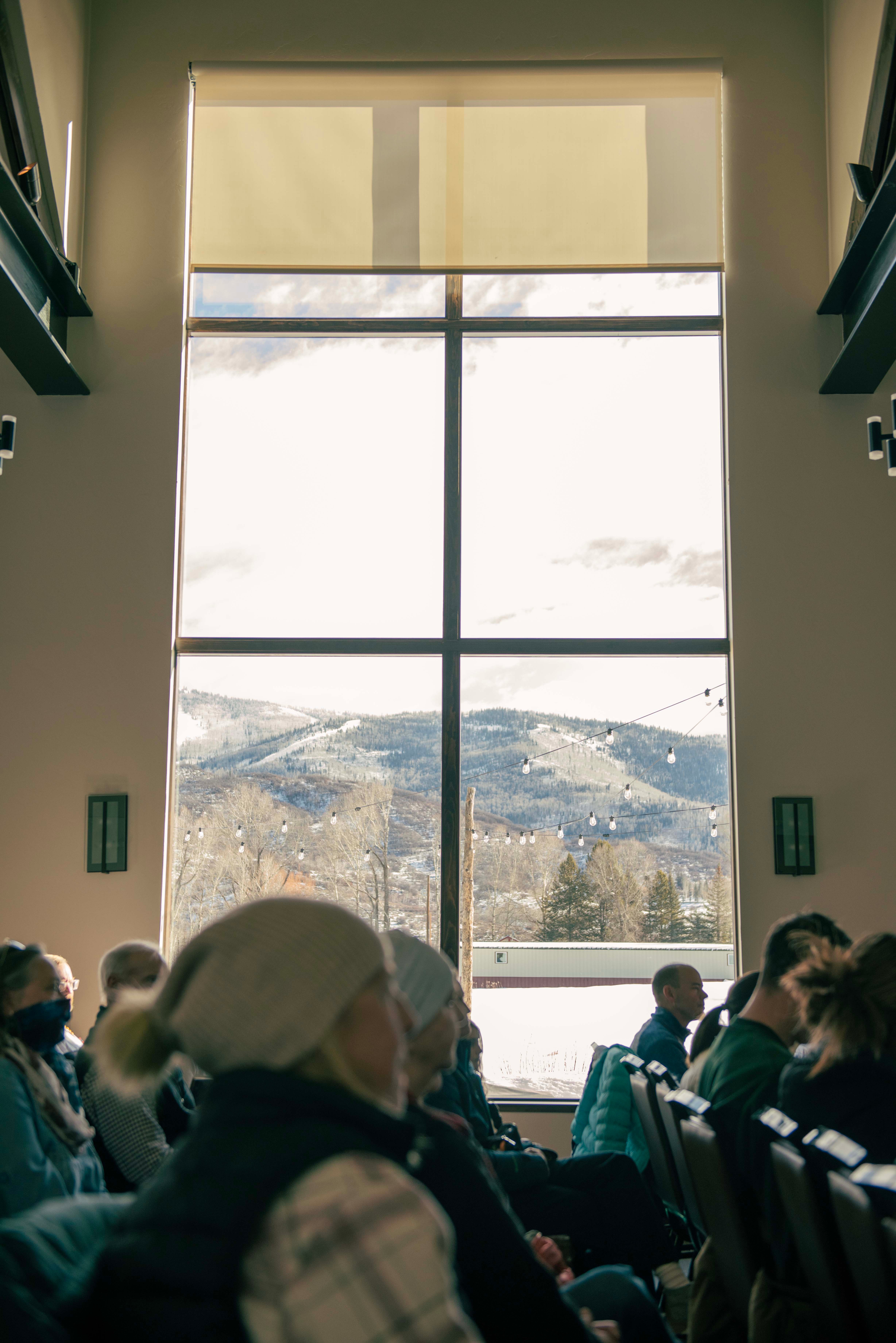 People seated indoors near large window with mountain view, some wearing masks, natural light, high ceiling, and curtains.