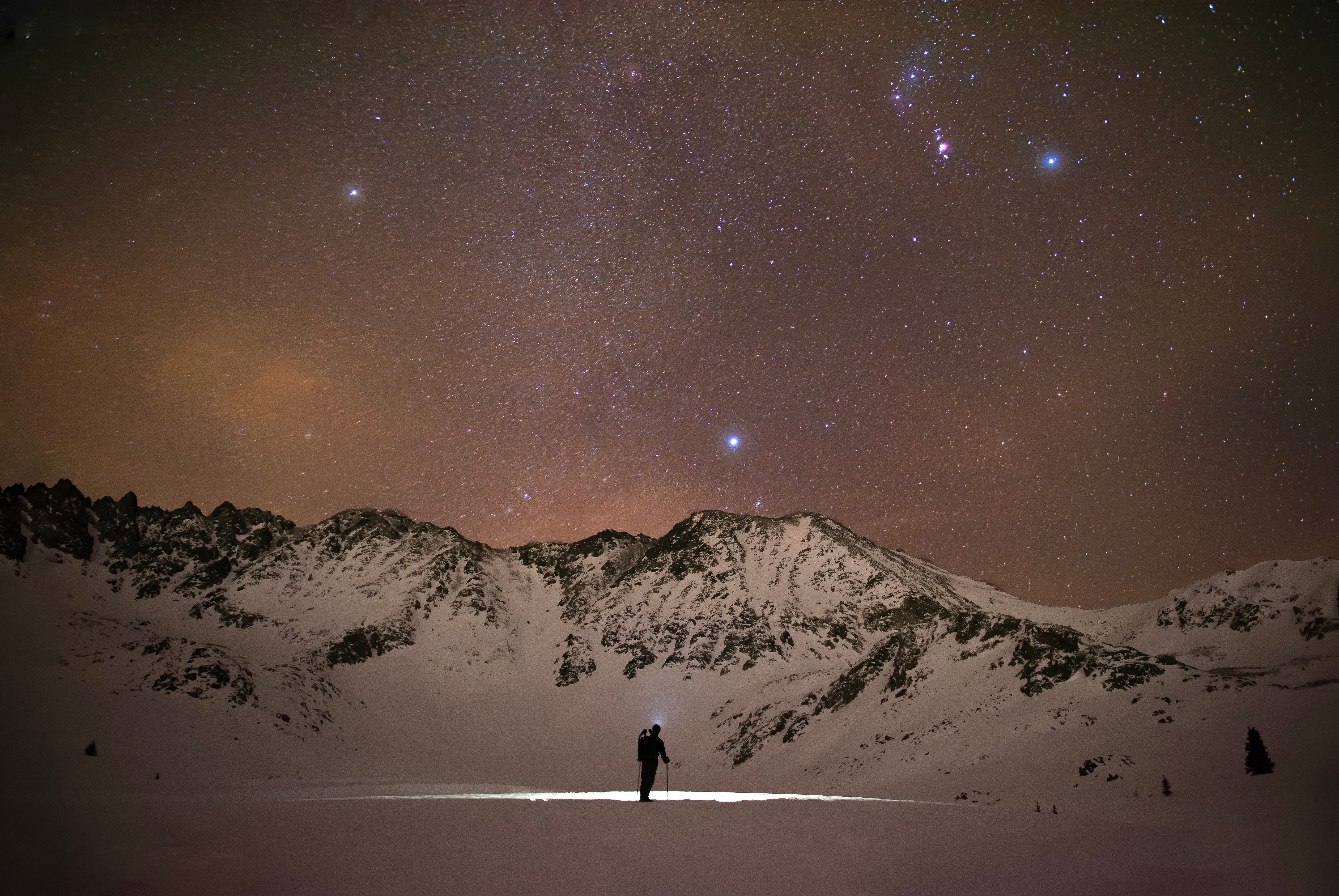 Person standing on snow-covered ground with mountains and starry sky in the background.