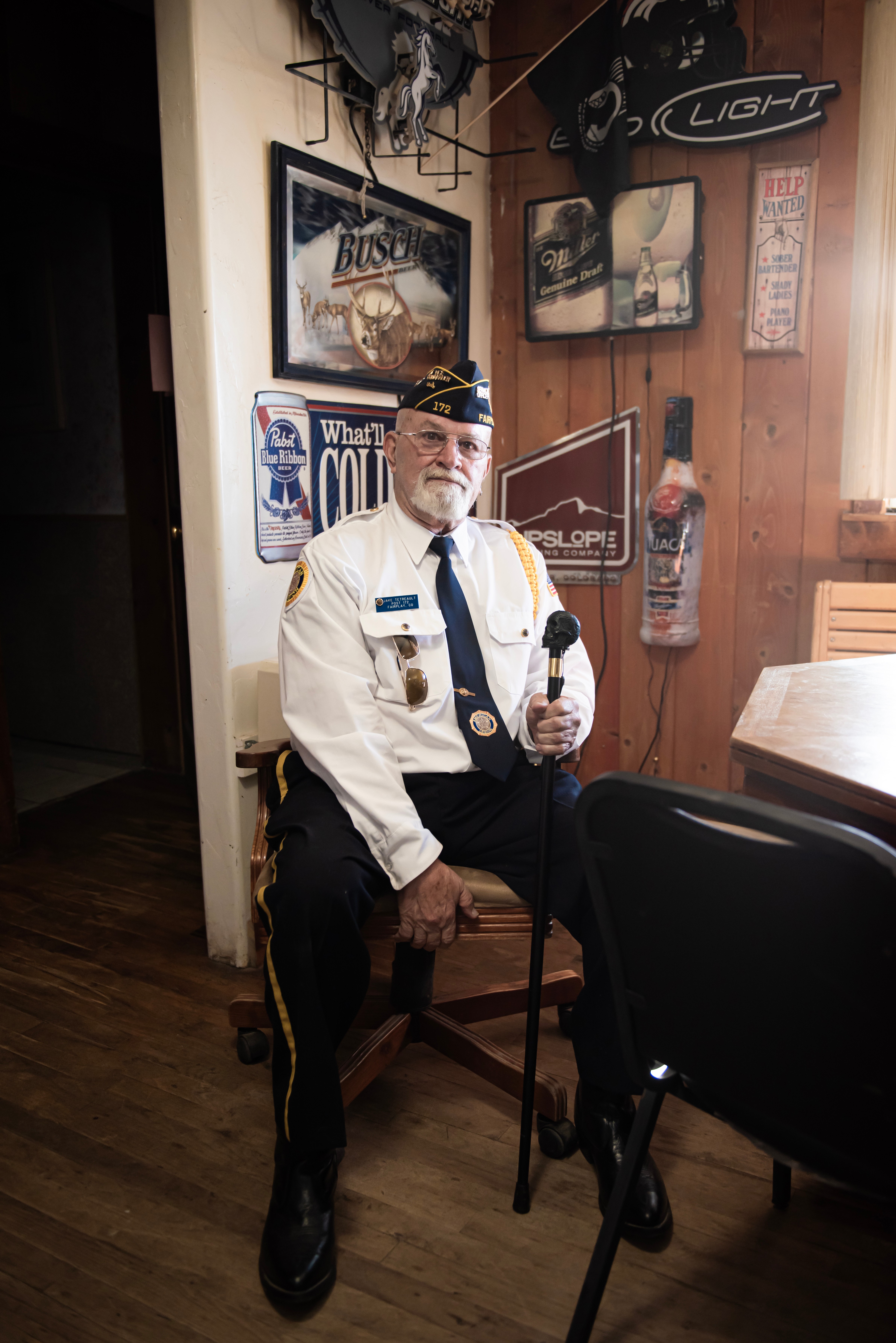 Older man in uniform sitting on a chair holding a bottle, in a room with wood-paneled walls and vintage signs.