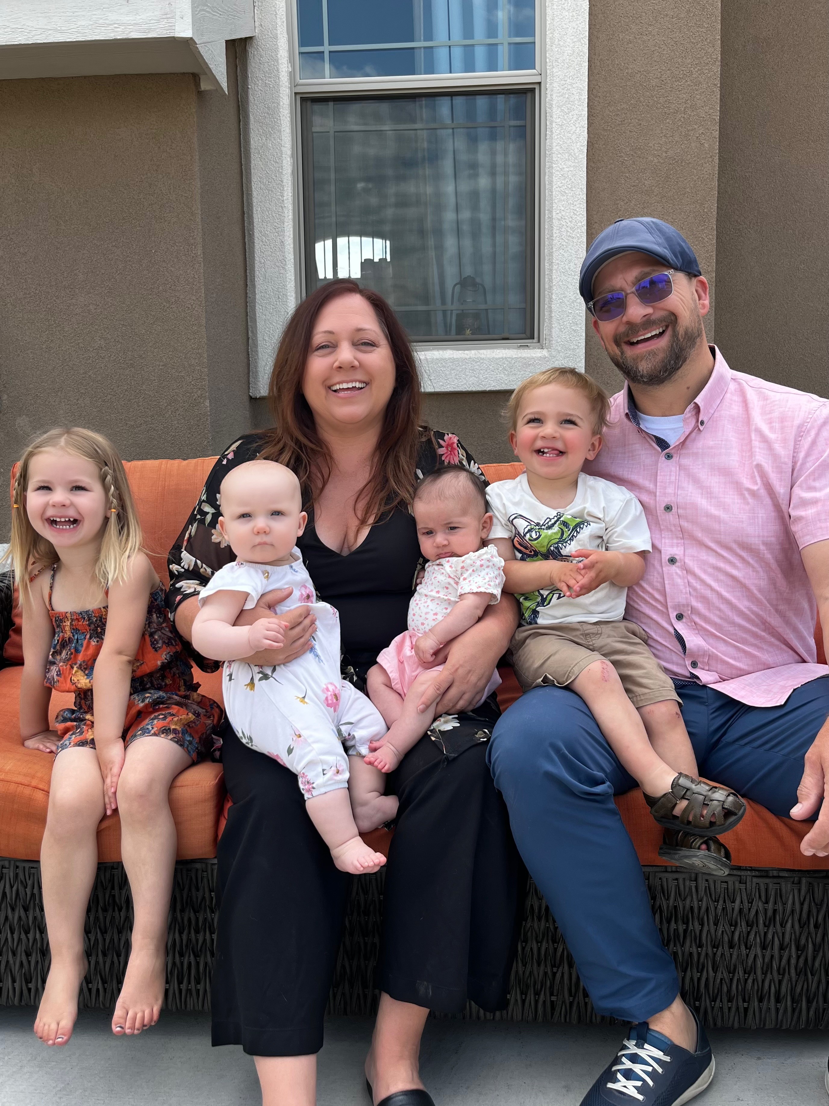 Family of six sitting on a bench outside, smiling, with a house and window in the background.