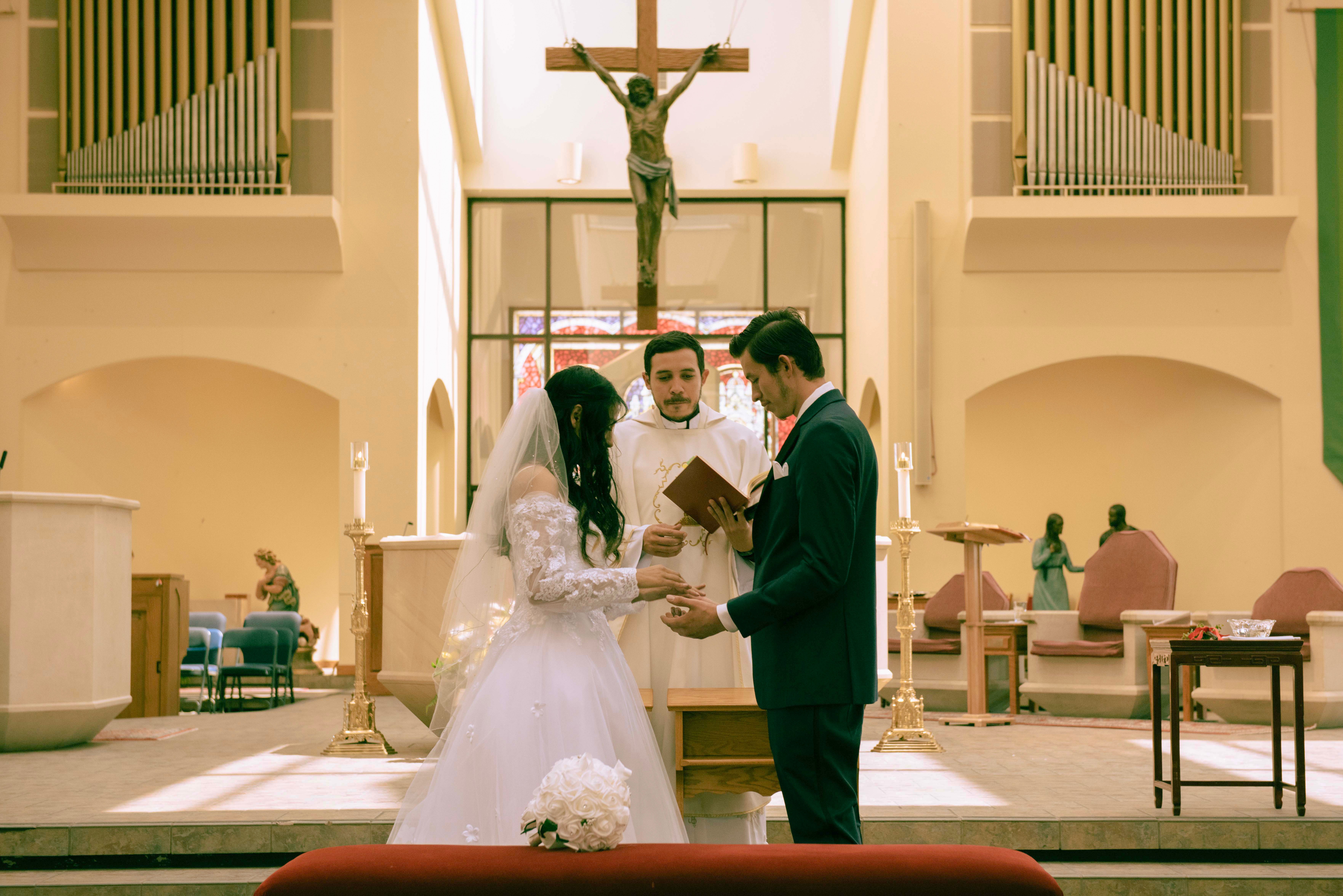 Bride and groom exchanging rings during wedding ceremony inside church with crucifix and altar in background.