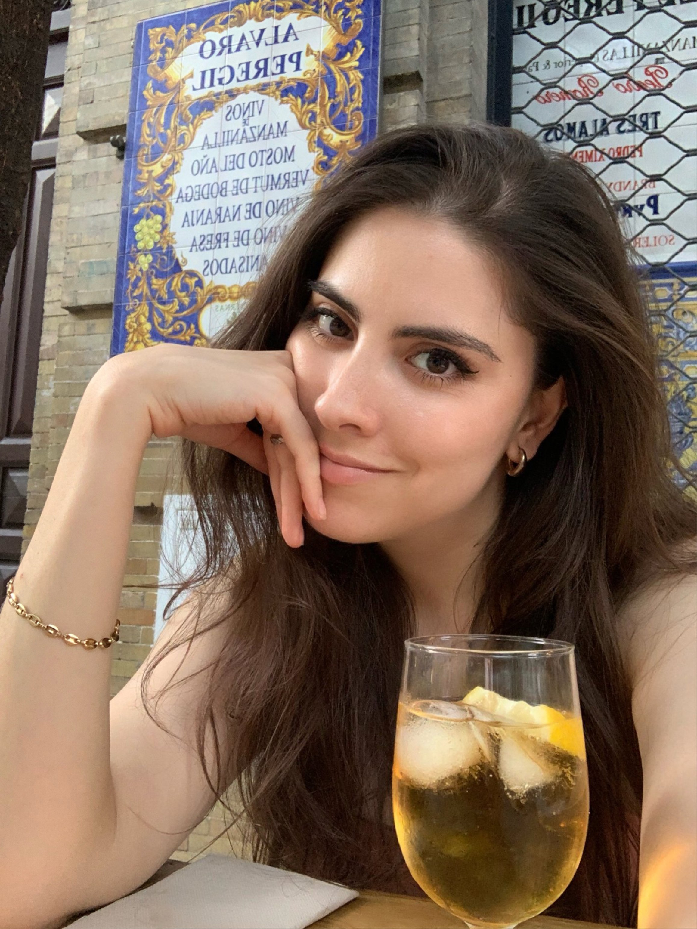Young woman with long brown hair sitting at a table, smiling, holding her chin, with a glass of drink in front of her, outdoors.
