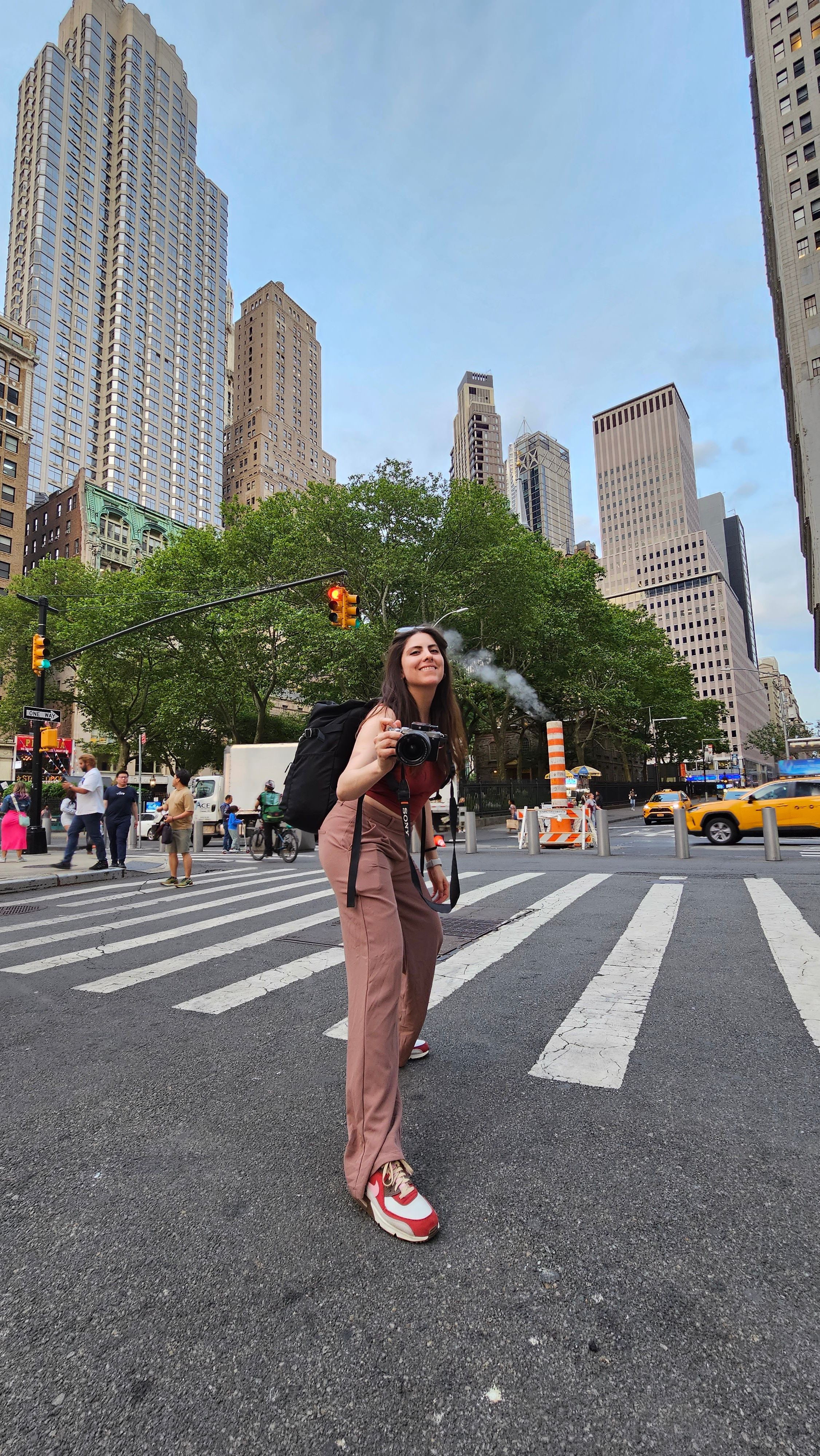 Person crossing a city street with tall buildings, trees, and yellow taxis in the background.