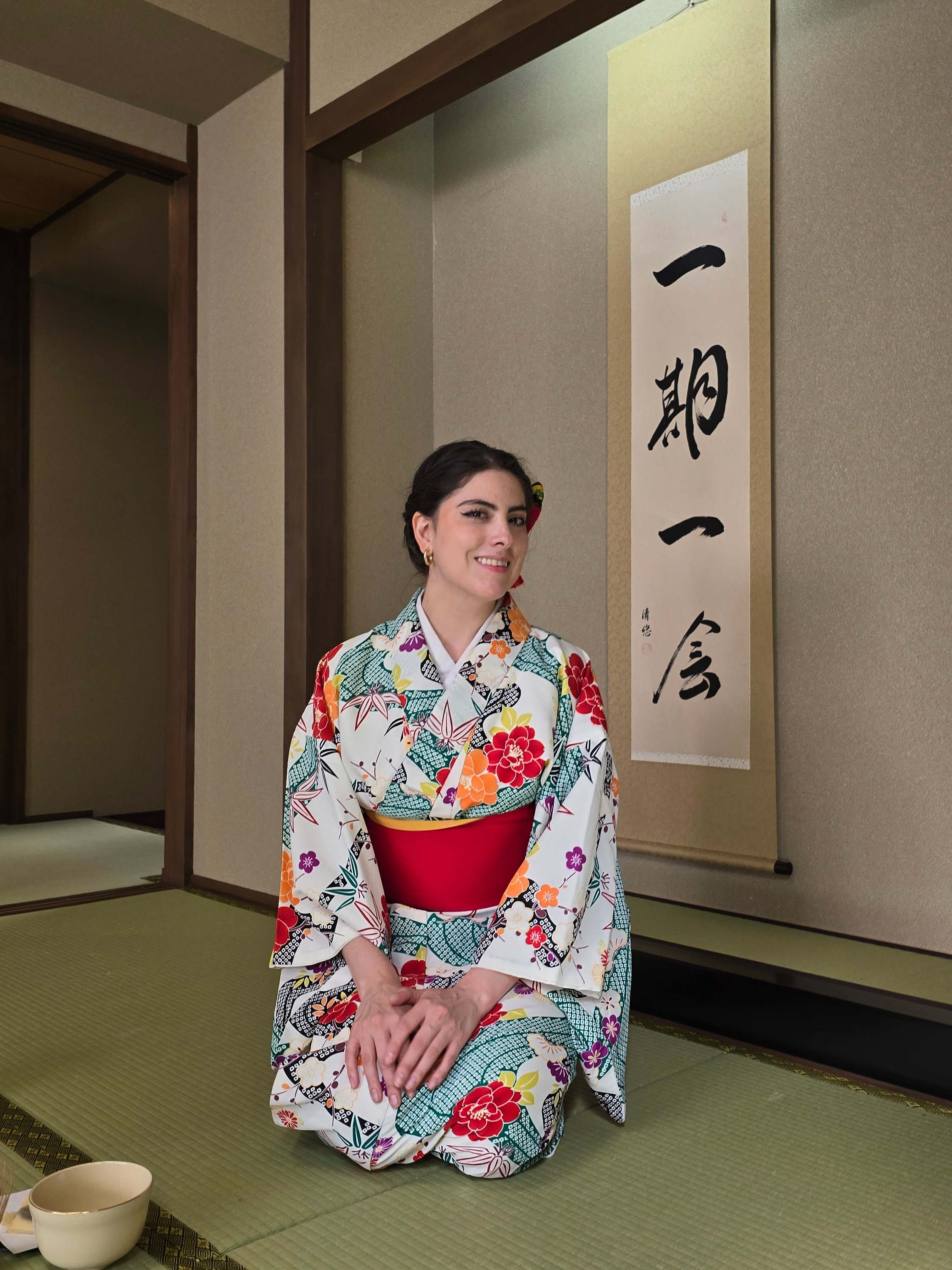 Woman in colorful kimono kneeling on tatami mat in traditional Japanese room with calligraphy scroll behind her.