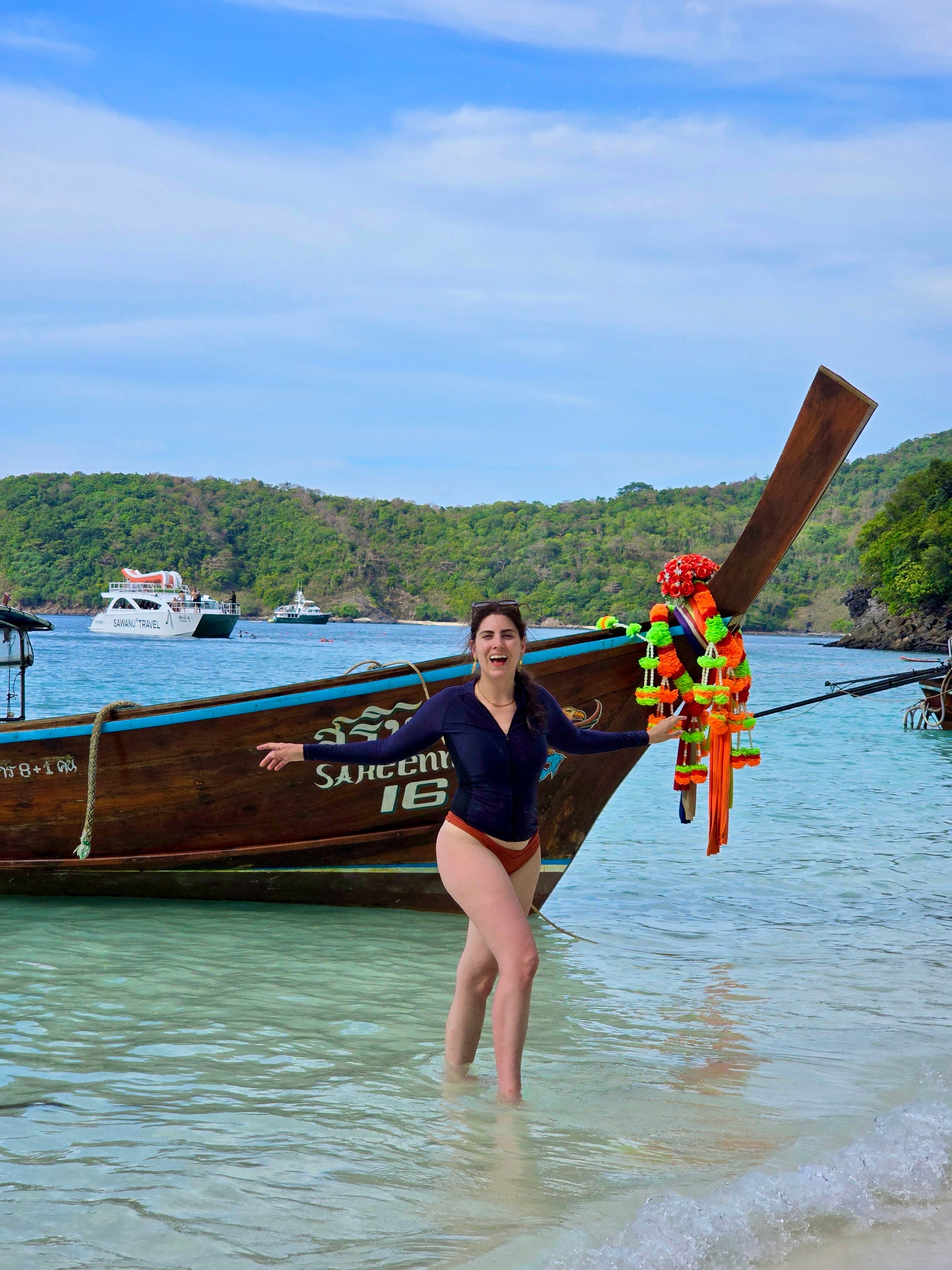 Woman standing in water near a boat, holding the boat's bow with a colorful decoration, with a river and green hills in background.