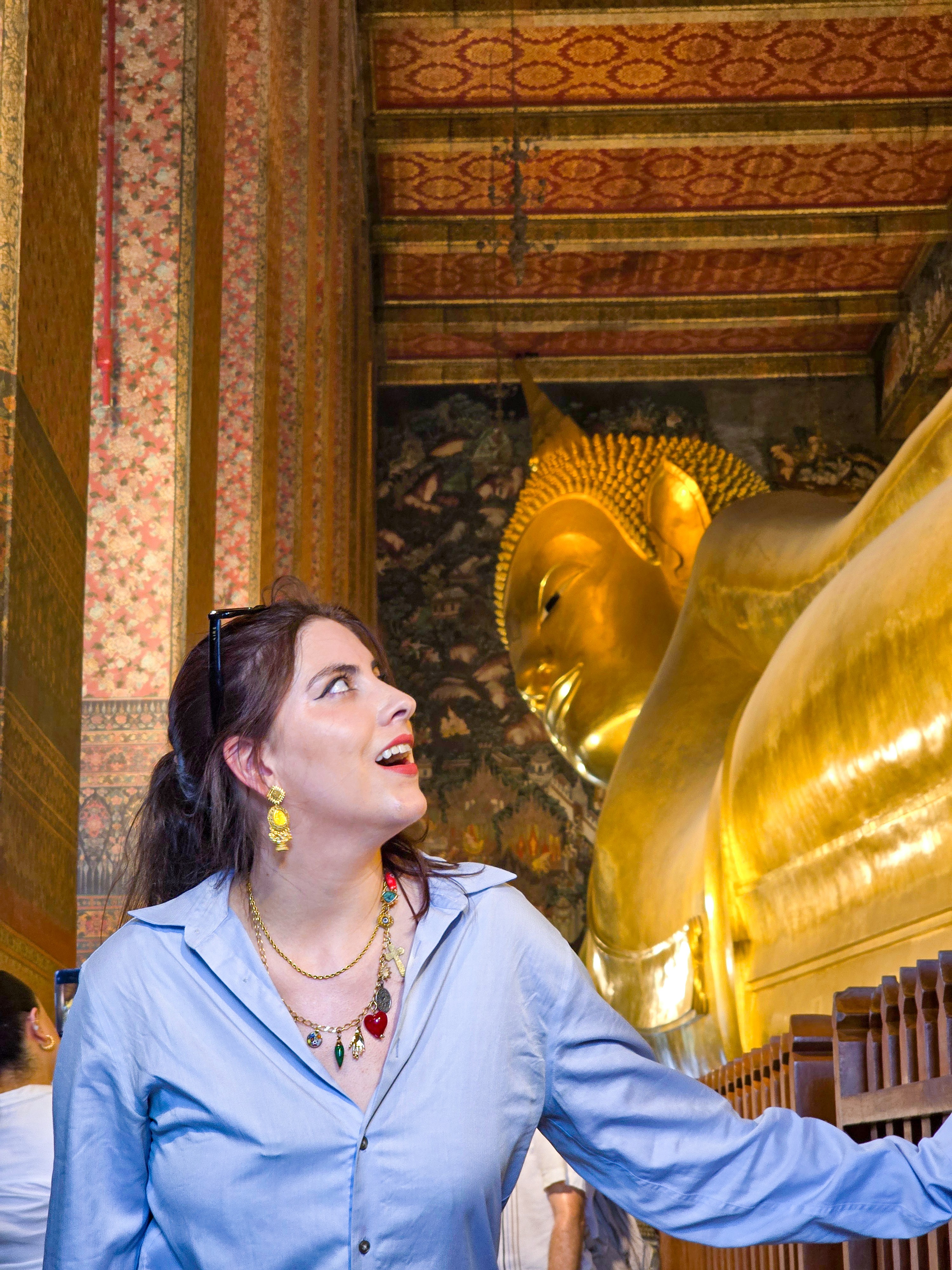 Woman with dark hair and colorful jewelry looks up at a large golden reclining Buddha inside a temple.