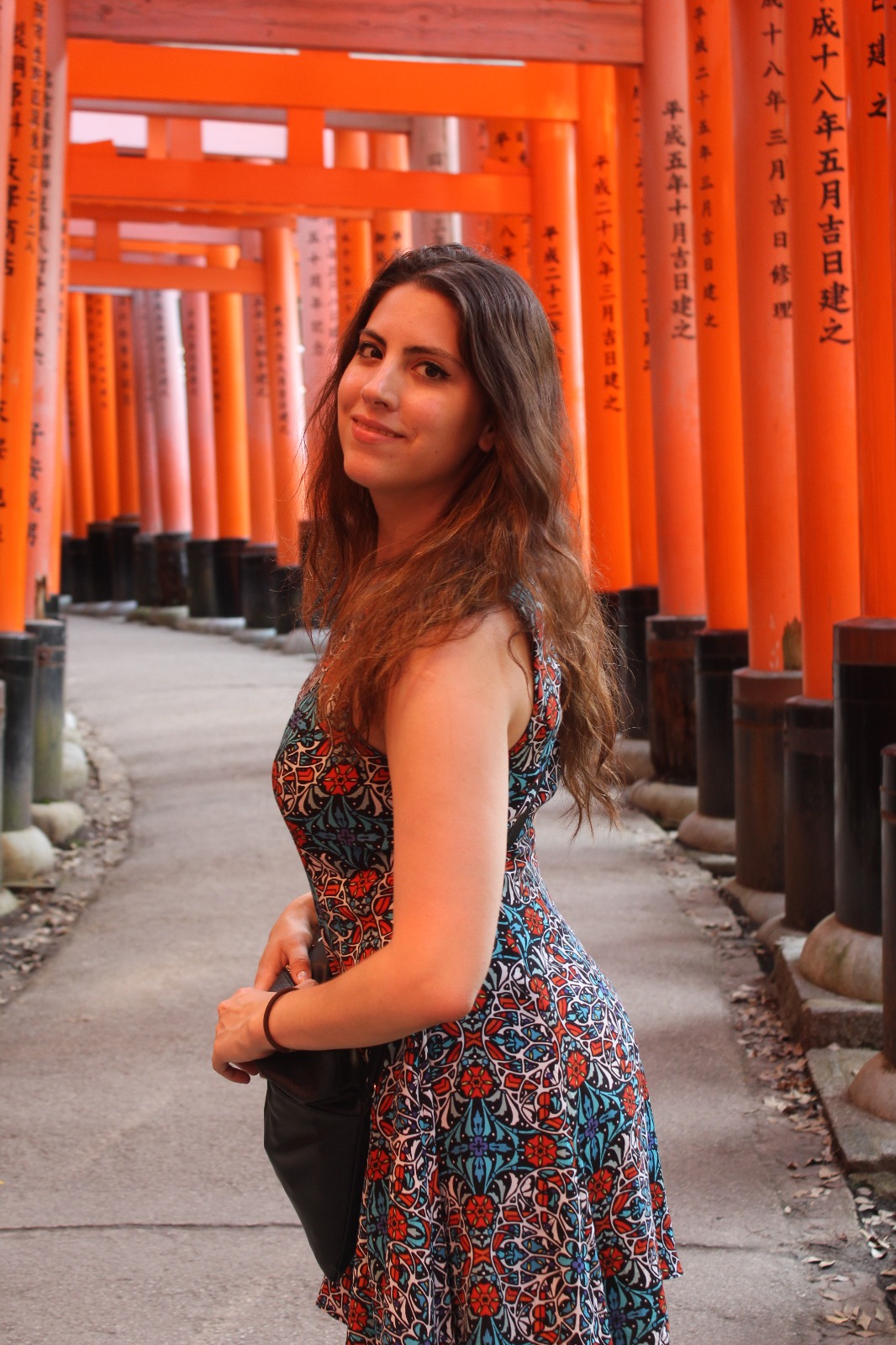 Young woman with long hair standing among orange torii gates at a shrine, smiling at the camera.