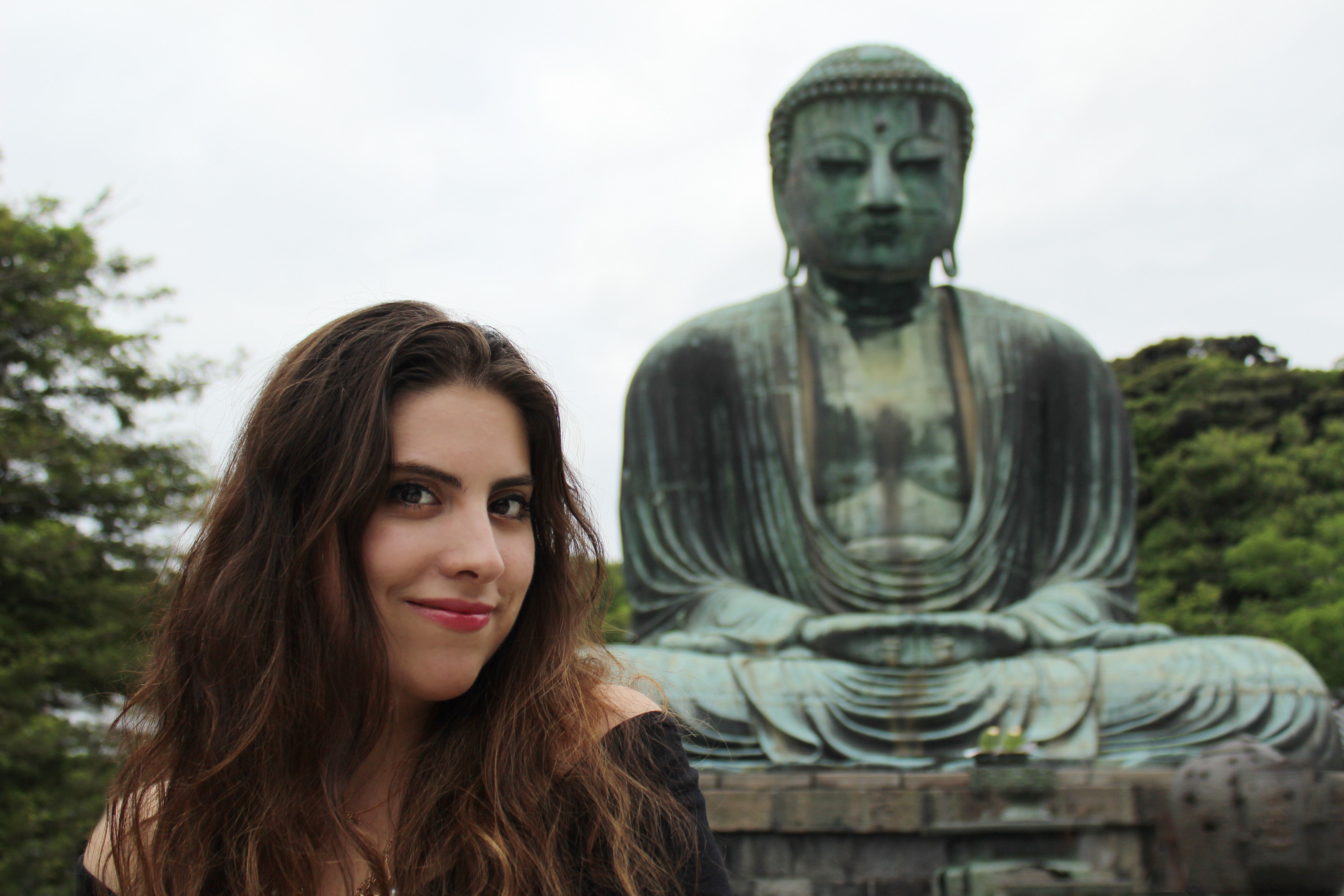 Woman with long brown hair smiling in front of a large seated Buddha statue outdoors.