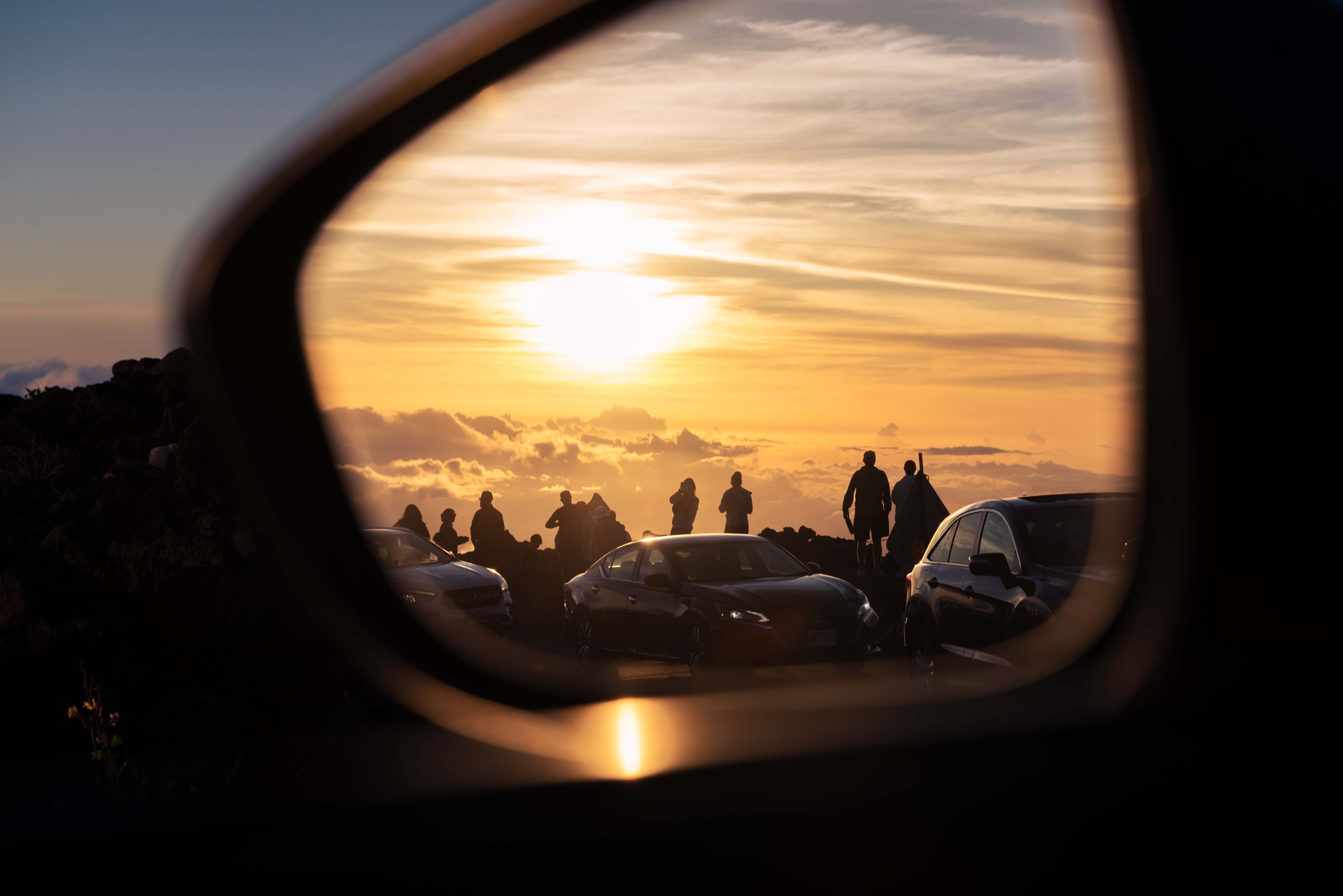 Sunset view through a car's side mirror with people and cars visible, warm colors in the sky.