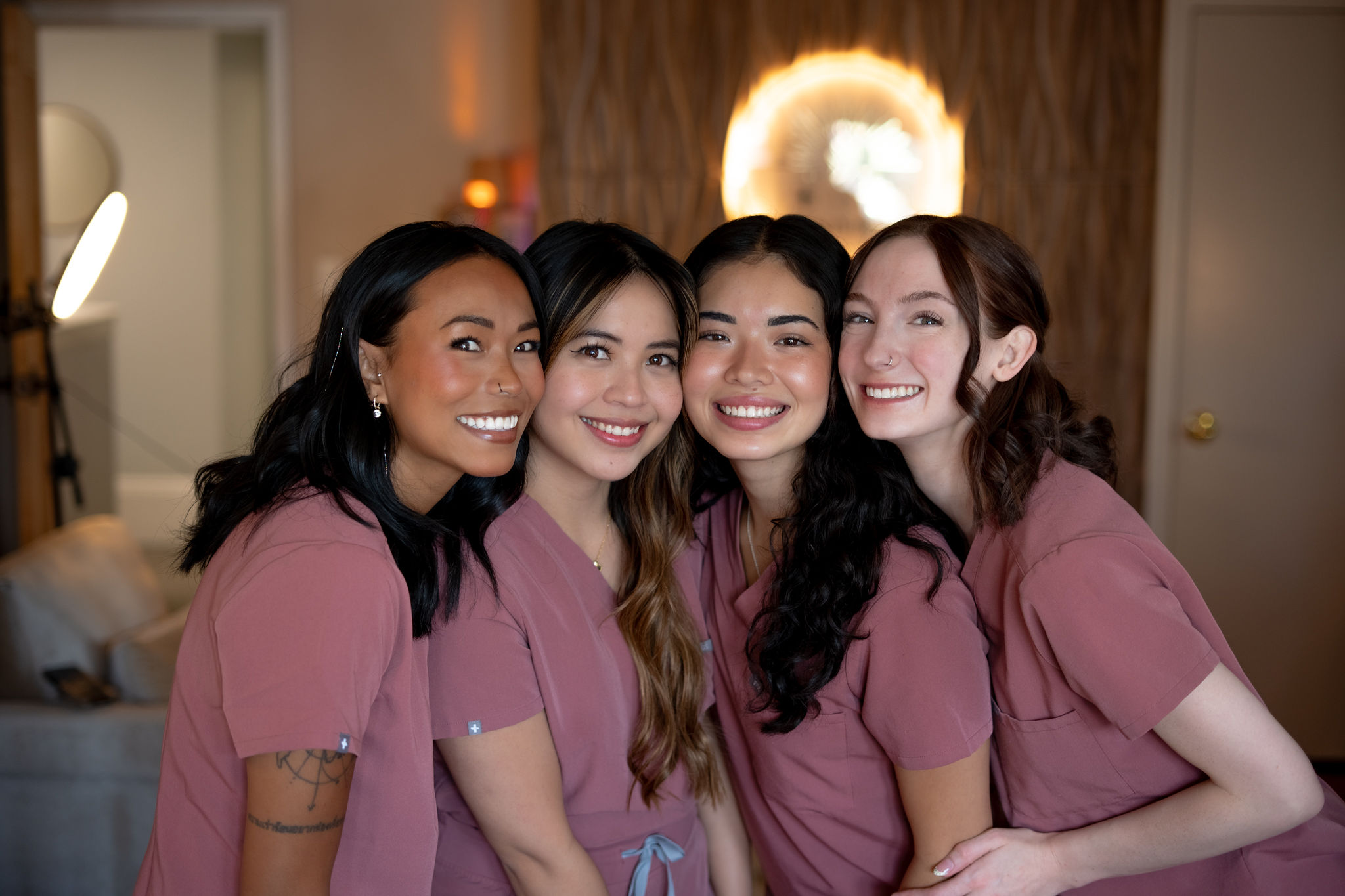 Four women in matching pink shirts smiling together indoors, with warm lighting and a circular light fixture in the background.