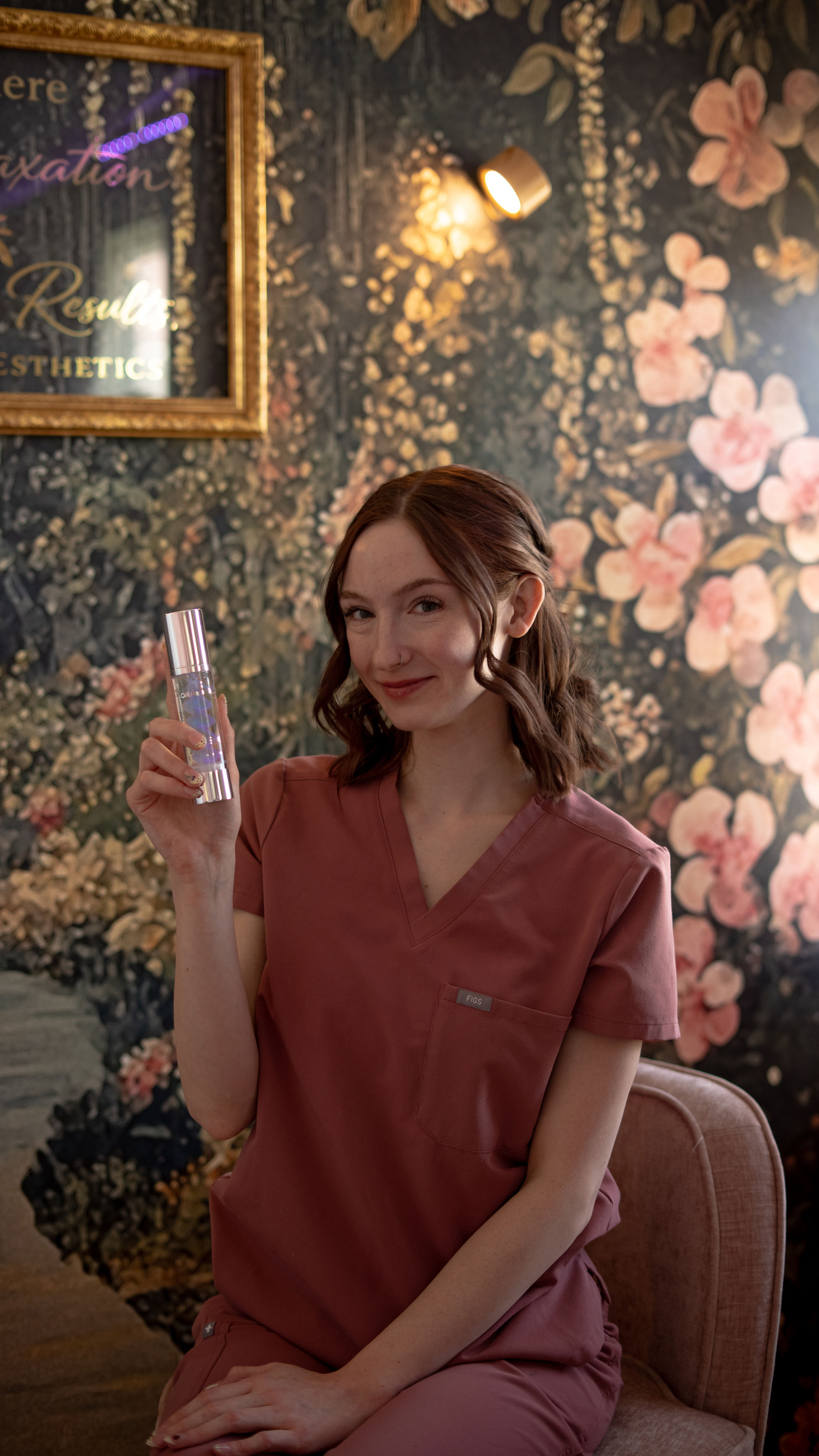 Young woman in pink scrubs sitting on a chair, holding a card, with floral wallpaper background.