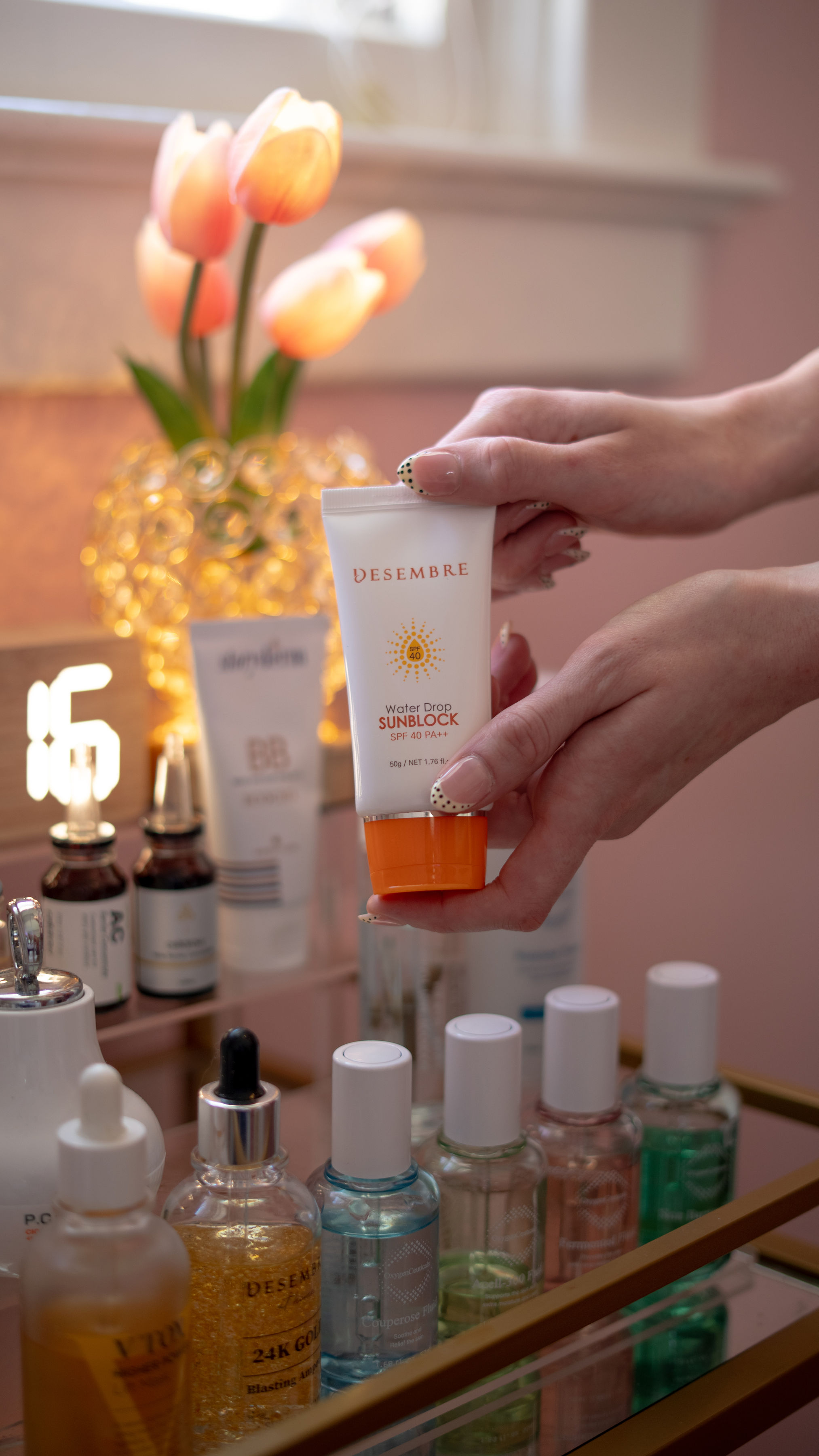 Person holding a skincare product with a background of tulips in a vase, various bottles on a table, and a pink wall.