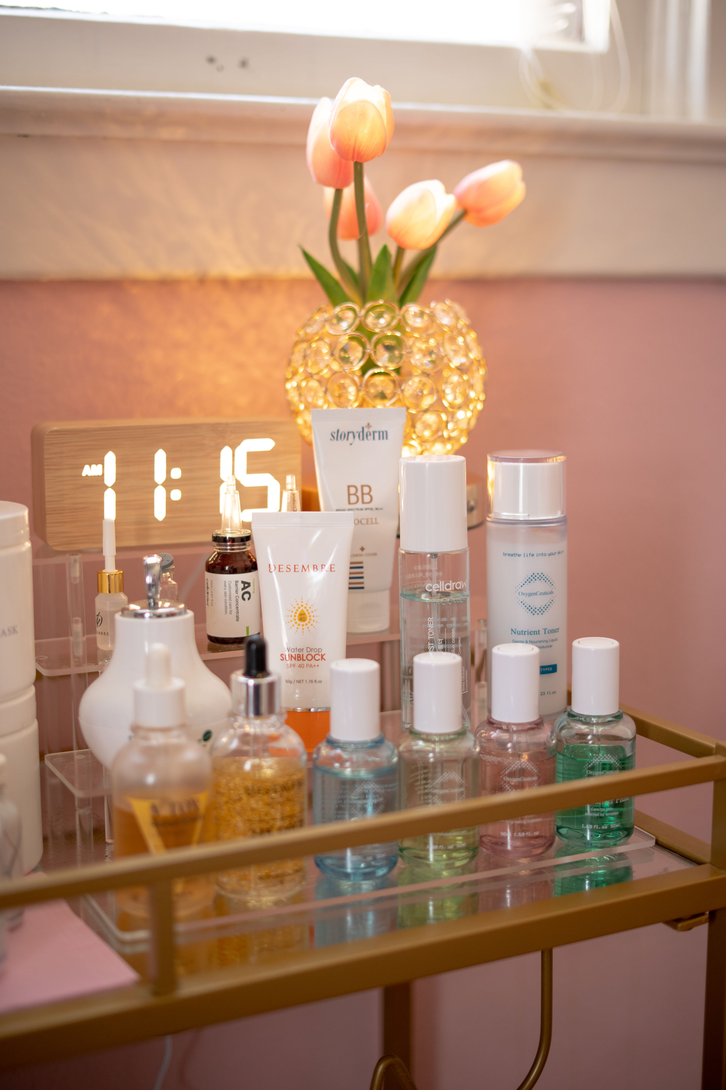 Pink and white tulips in a decorative glass vase on a table with skincare products, a digital clock showing 11:55, and a window in the background.