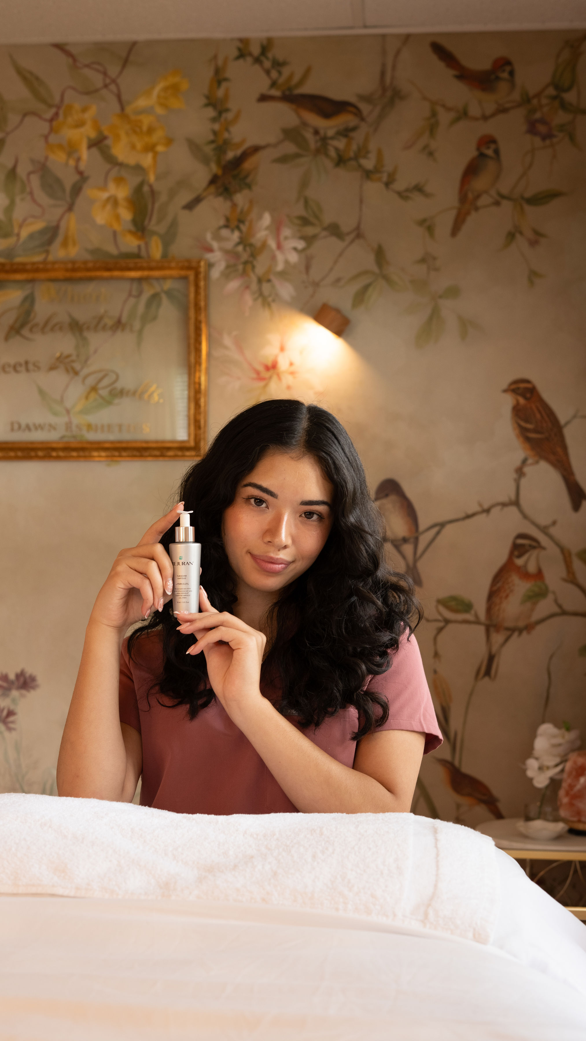 Young woman with dark curly hair holding a skincare product, sitting at a table in a decorated room.