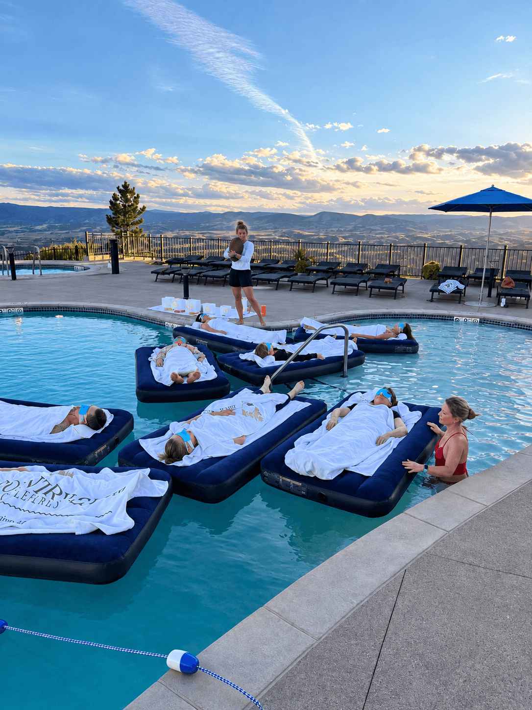 People relaxing on pool floats with towels, poolside chairs, and a person standing near the pool, with a scenic landscape and sky in the background.