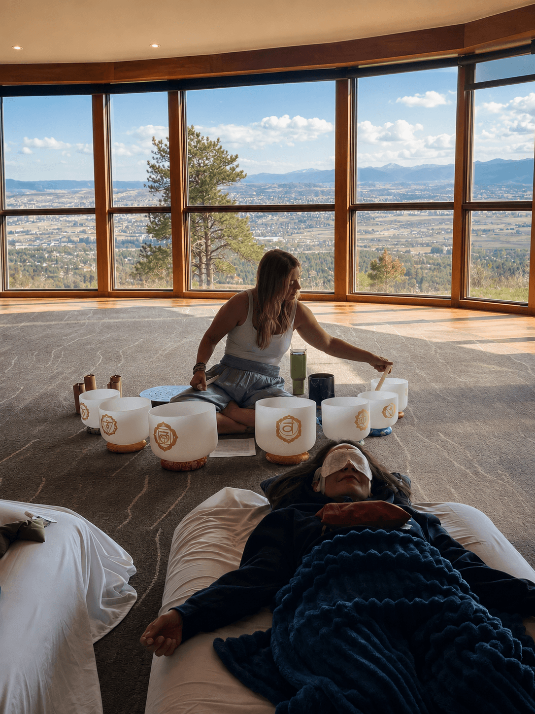 Person sitting on the floor with a woman, surrounded by singing bowls, in a room with large windows showing a landscape.