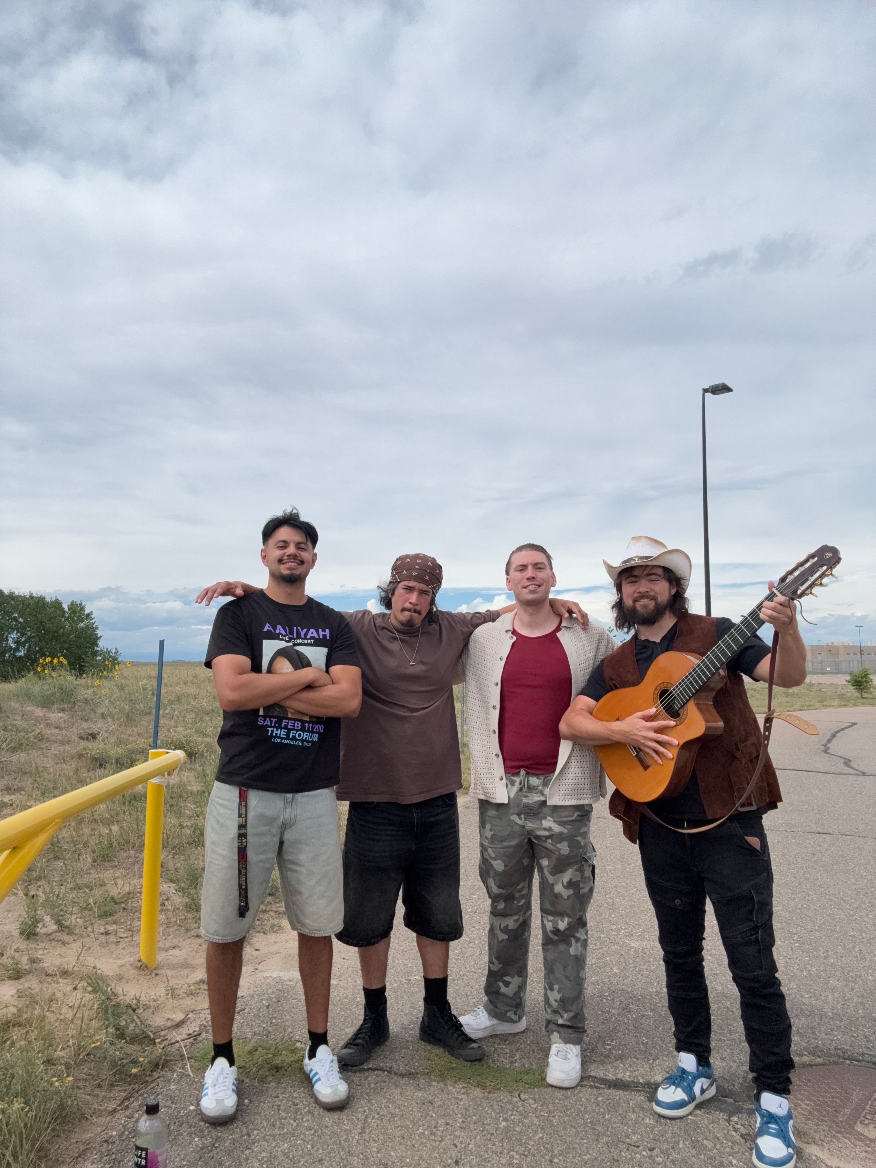 Four people standing outdoors, one playing guitar, others posing, cloudy sky in background.