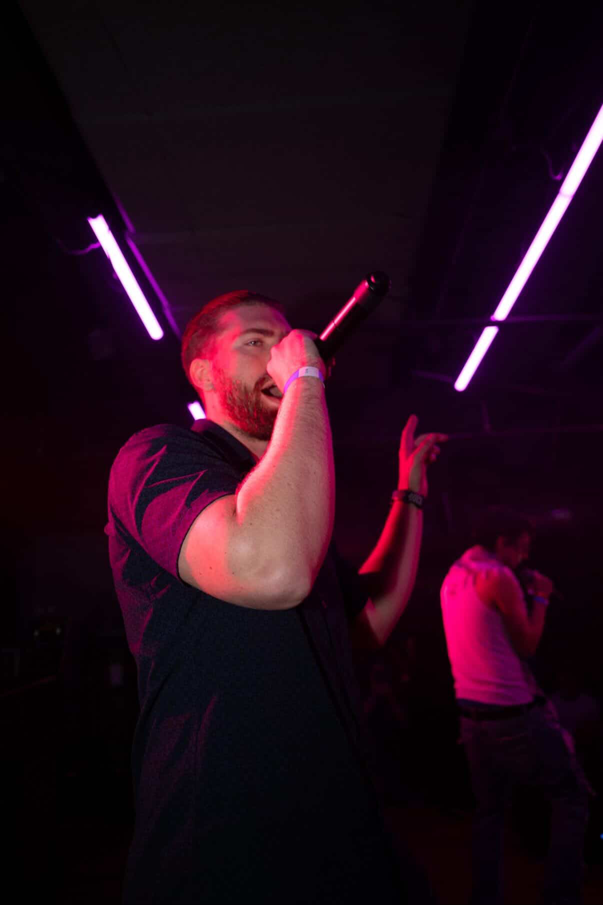 Person singing into a microphone on stage with purple lighting, dark background, and another person in the background.