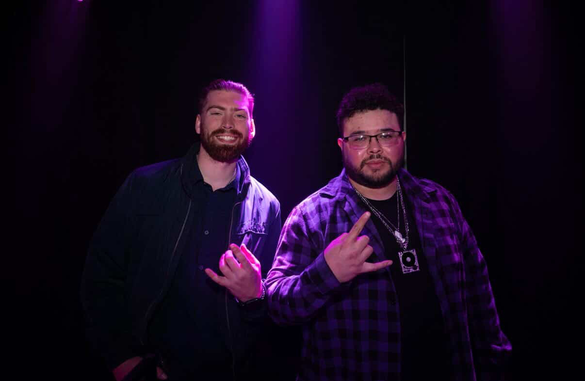 Two men standing on a stage with purple lighting, smiling, one making a hand gesture, the other holding a camera.