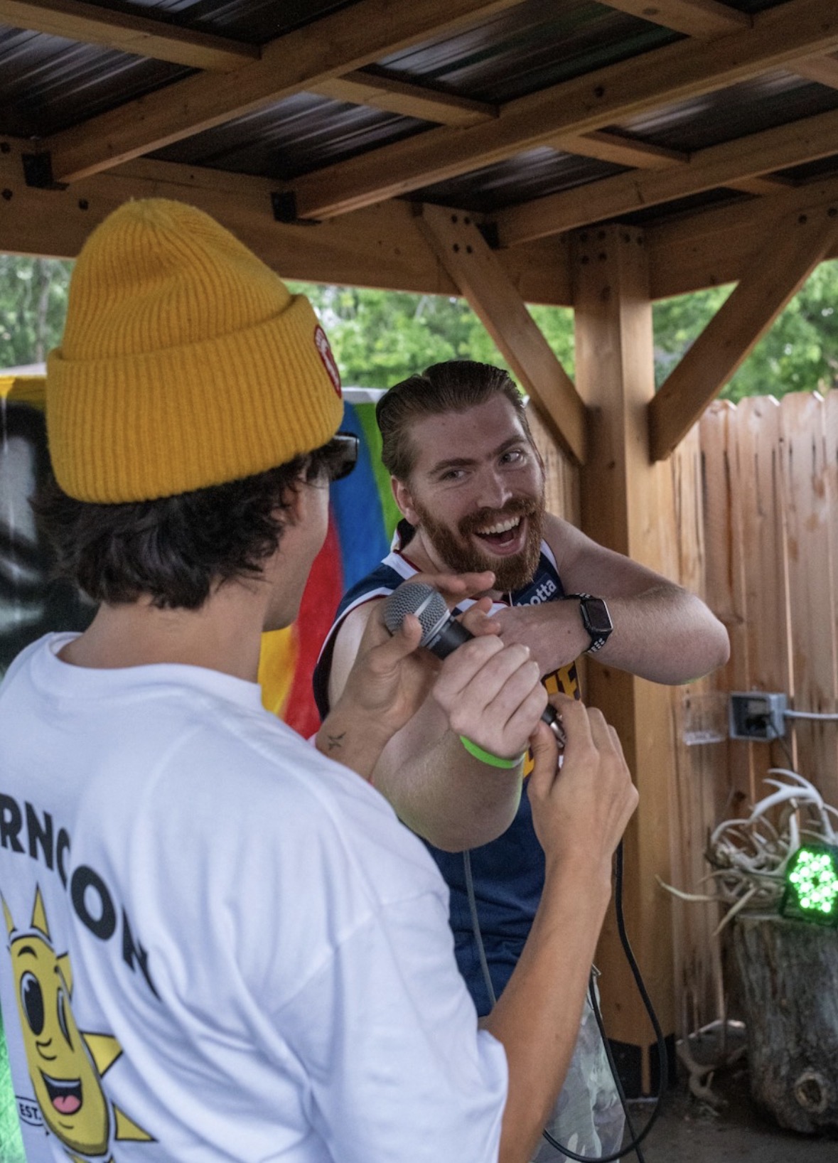 Two men talking outdoors under a wooden roof, one wearing a yellow beanie, smiling and gesturing with hands.