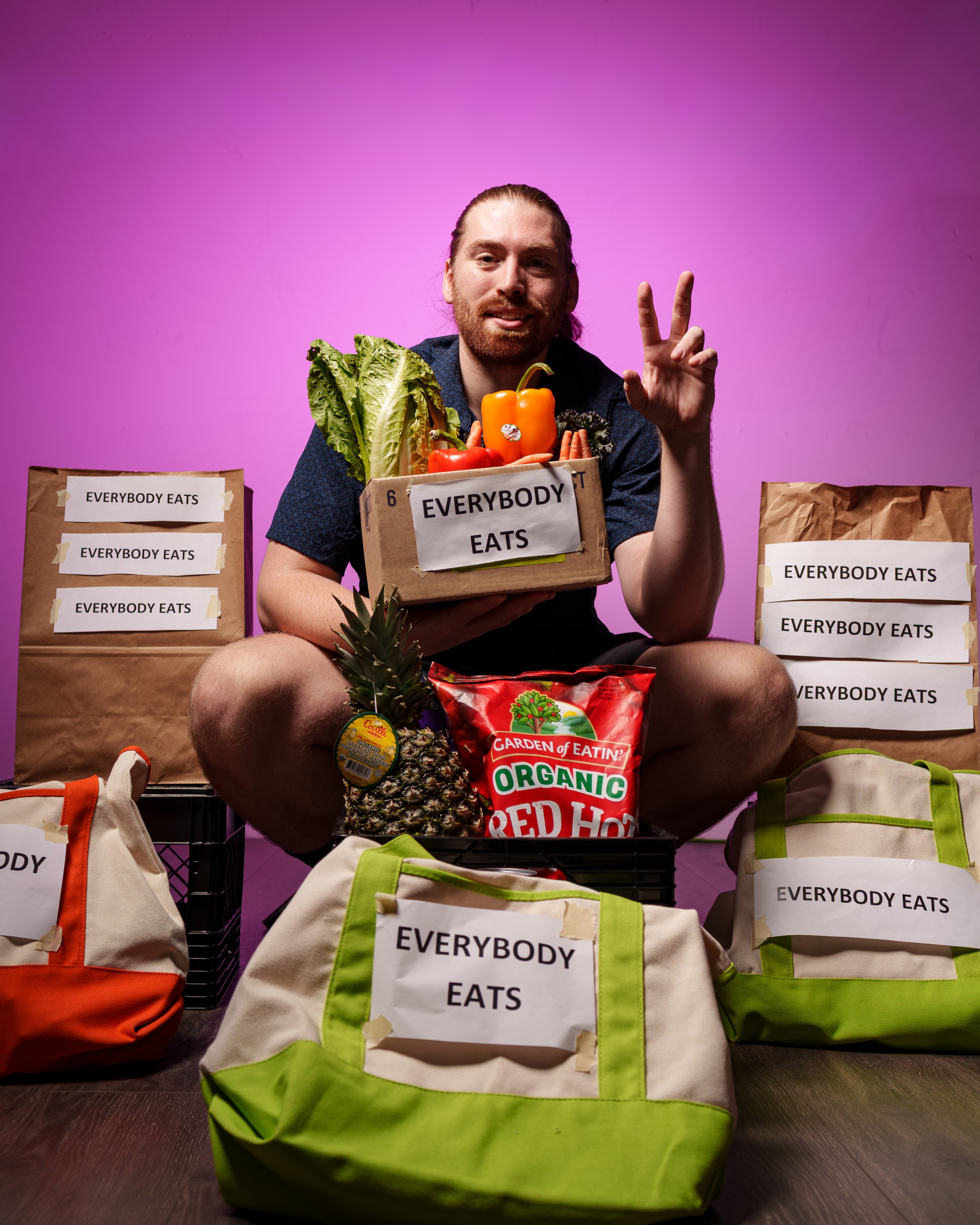 Man sitting surrounded by grocery bags, holding a box of vegetables, making a peace sign, with a purple background.
