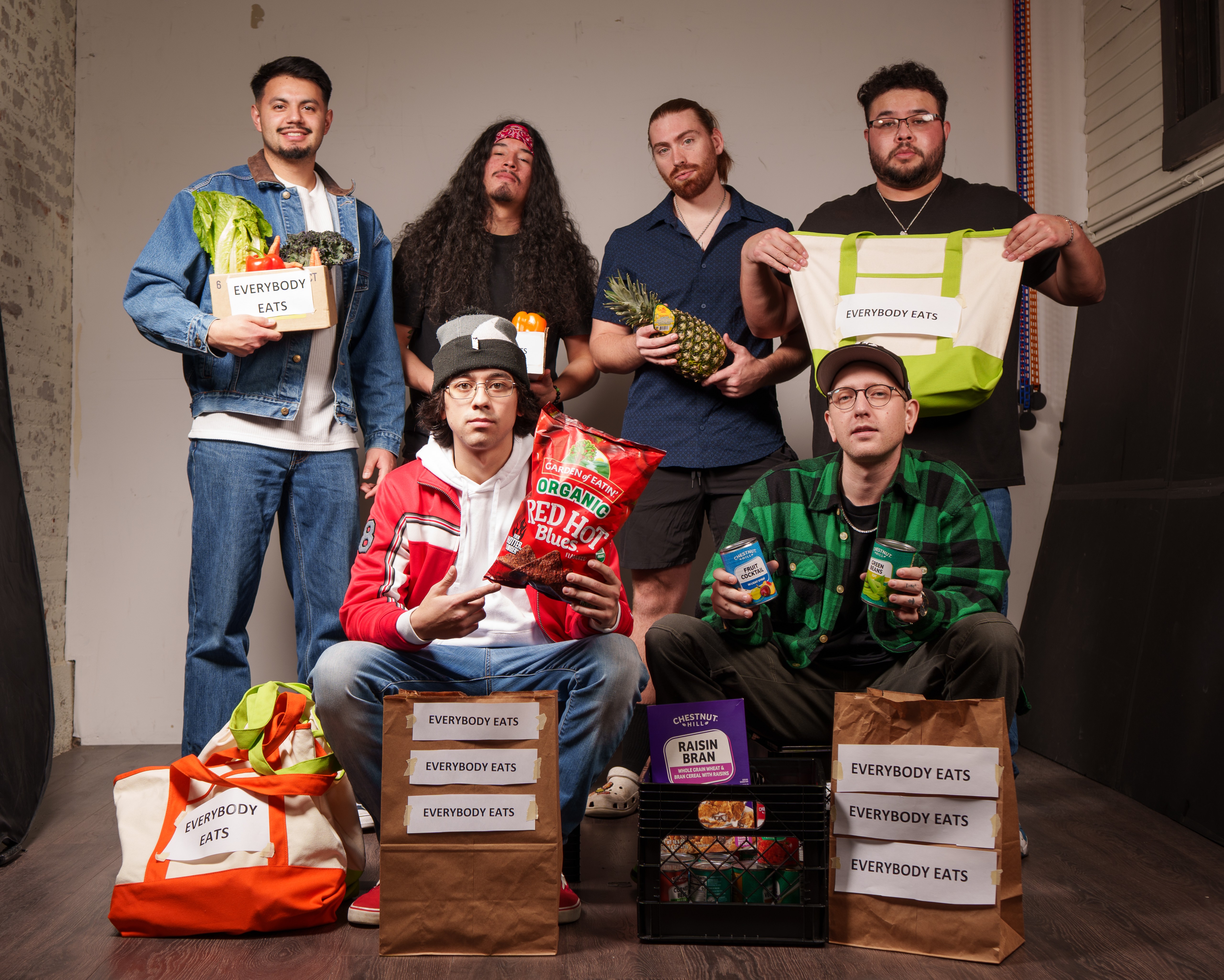 Six people pose with food and grocery bags, some holding snacks and drinks, indoors against a plain wall.