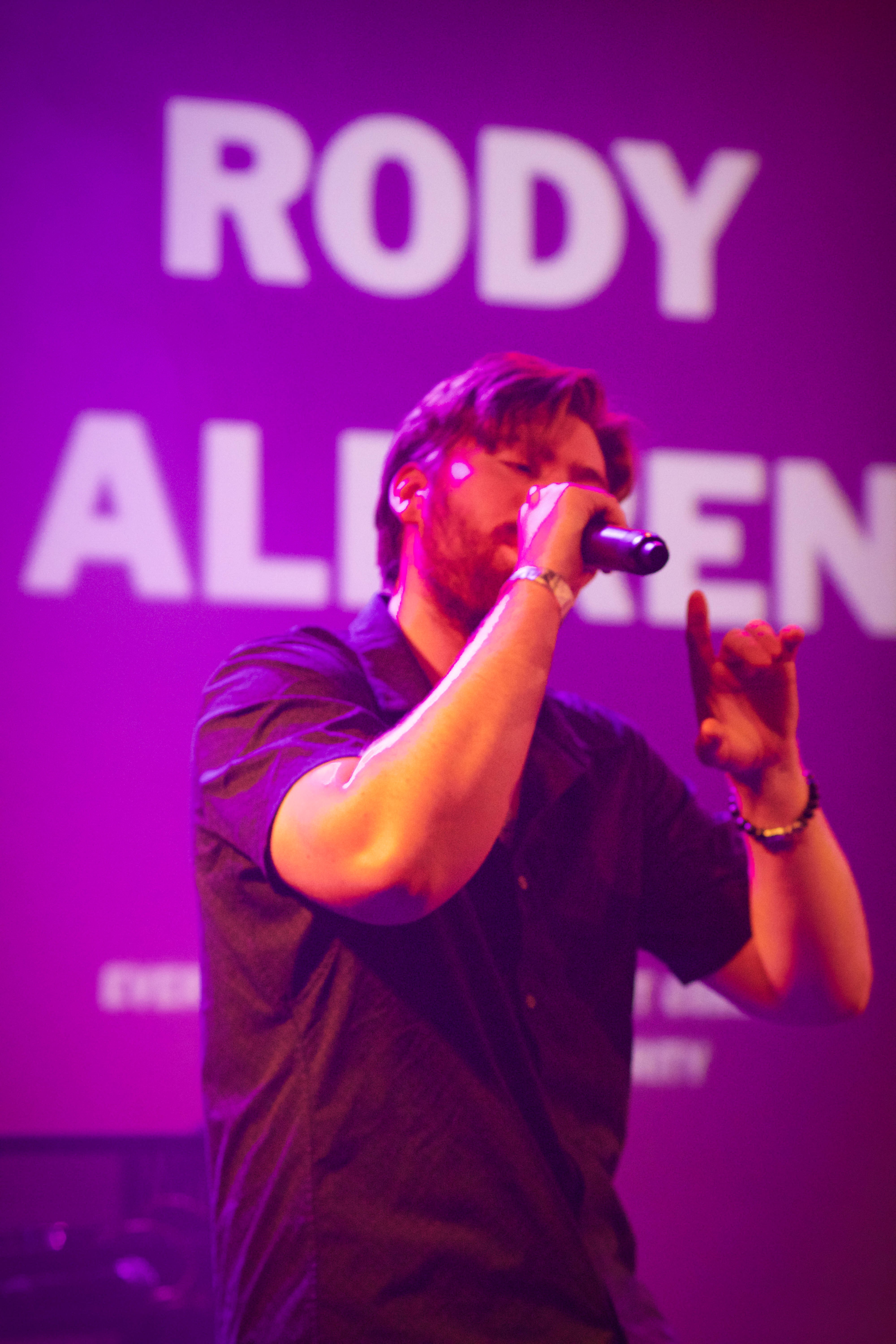 Person singing into microphone on stage with purple background and large text, wearing glasses and dark shirt.
