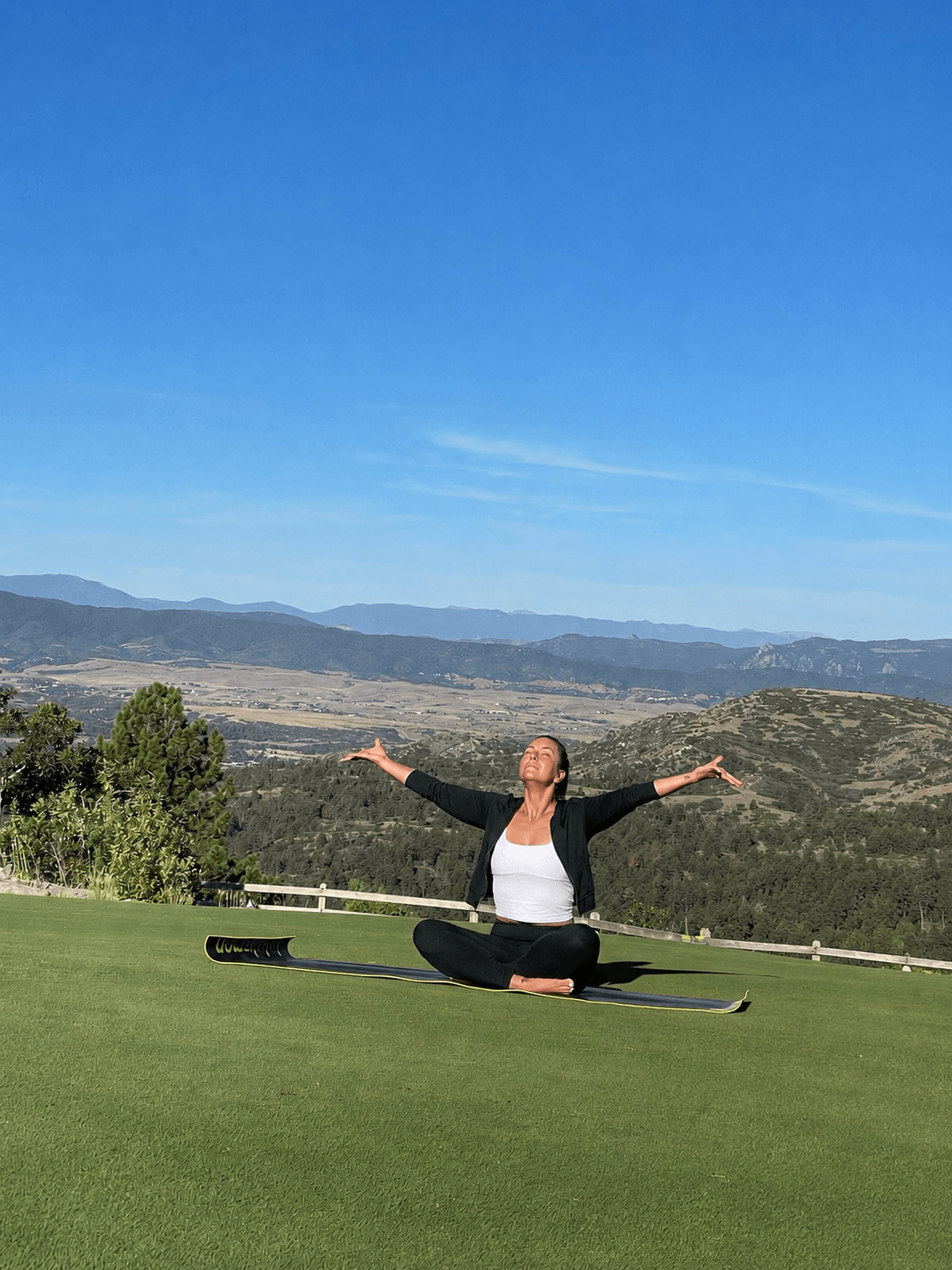 Person sitting cross-legged on grass with arms outstretched, outdoors with hills and blue sky in background.