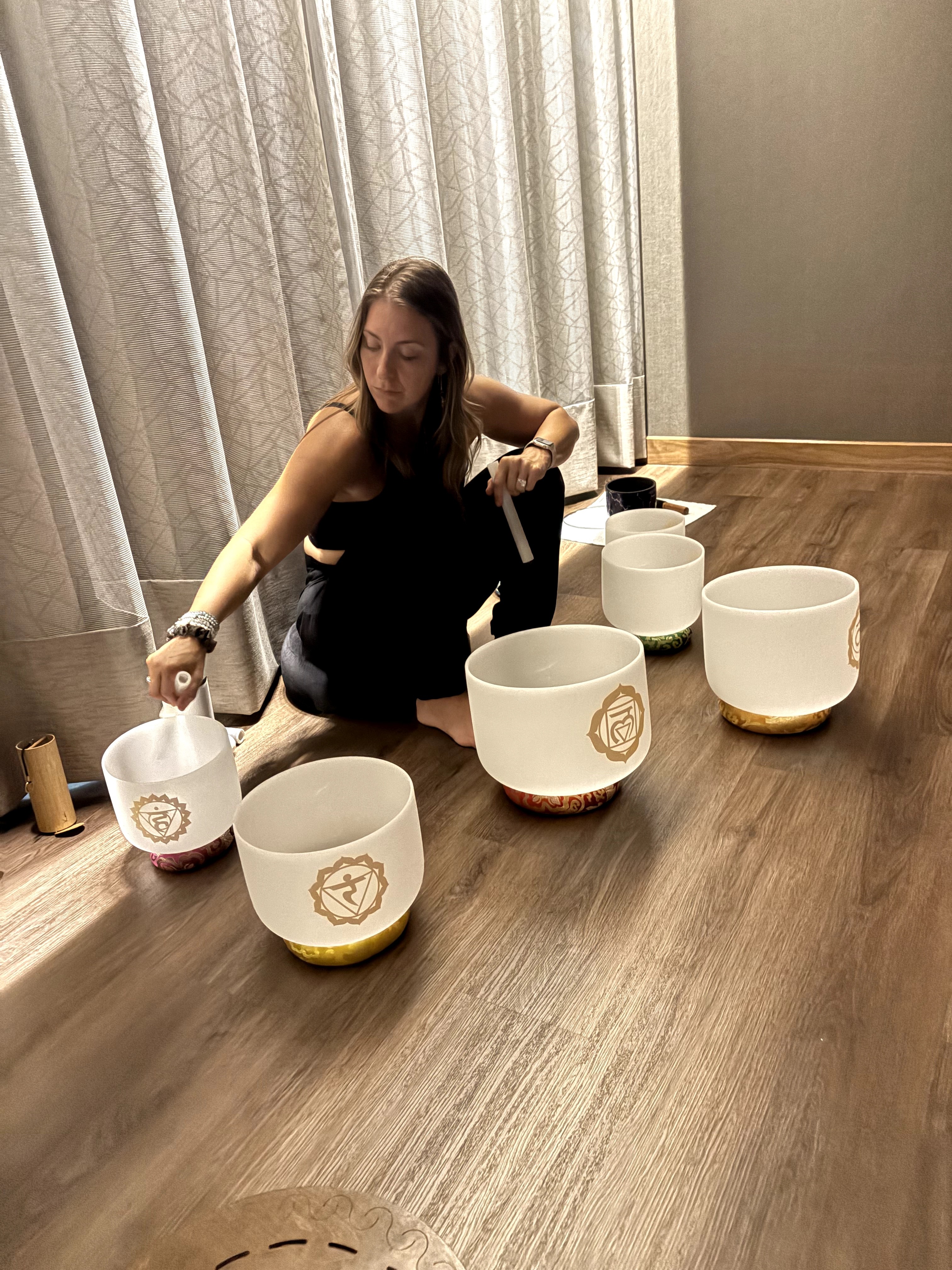 Woman playing singing bowls on a wooden table with curtains in the background.