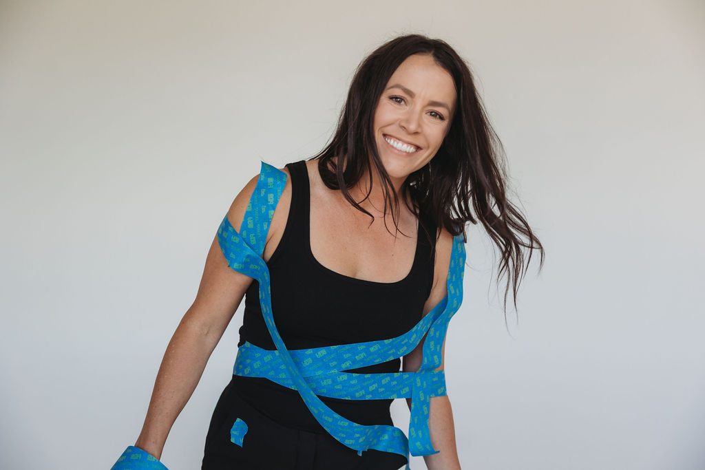 Smiling woman with long dark hair wearing a black top and blue patterned wrap, standing against a plain background.