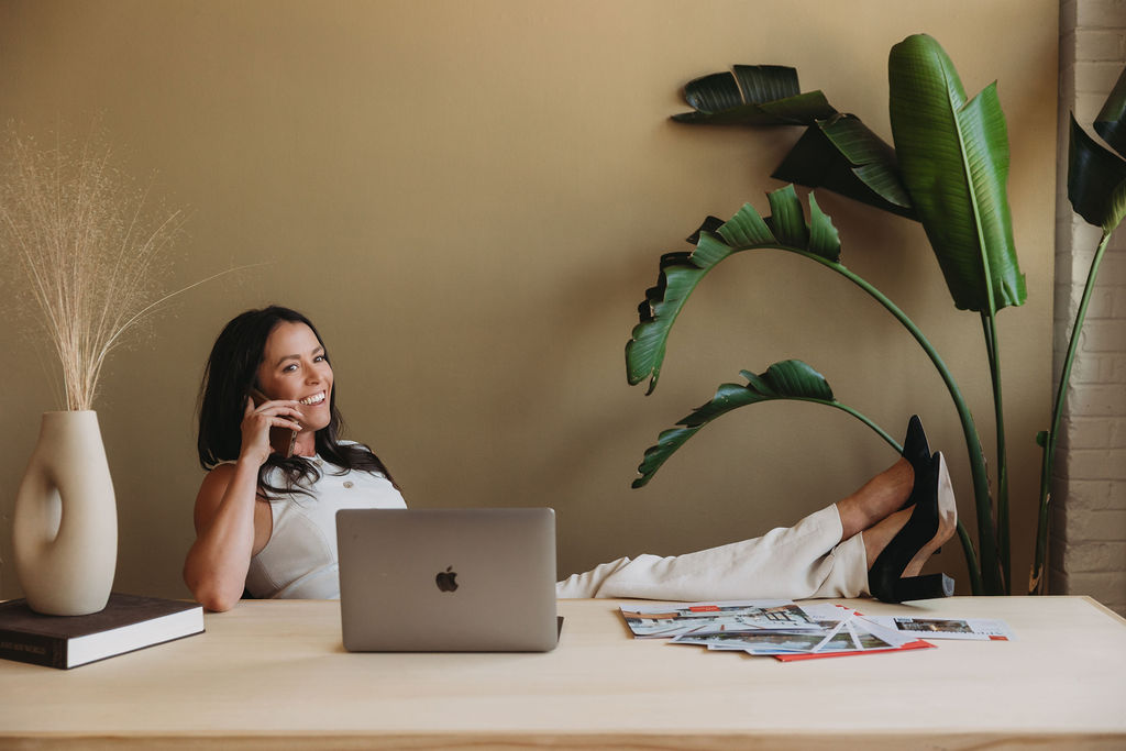 Woman lying on a desk with legs extended, using a laptop, surrounded by plants and magazines, in a room with beige walls.