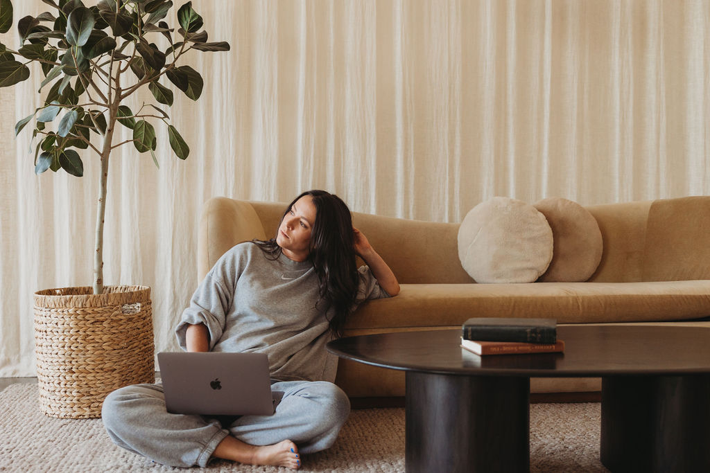Person sitting on the floor with a laptop, leaning against a sofa in a room with curtains and a potted plant.