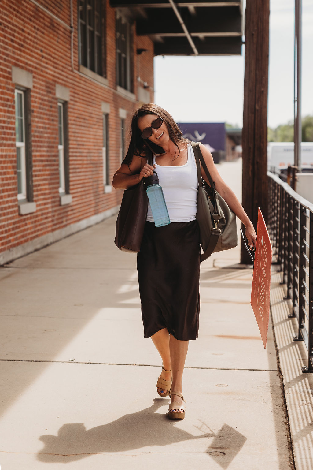 Woman walking outdoors with a bag, sunglasses, and an umbrella, beside a brick building and railing, sunlight casting shadow.
