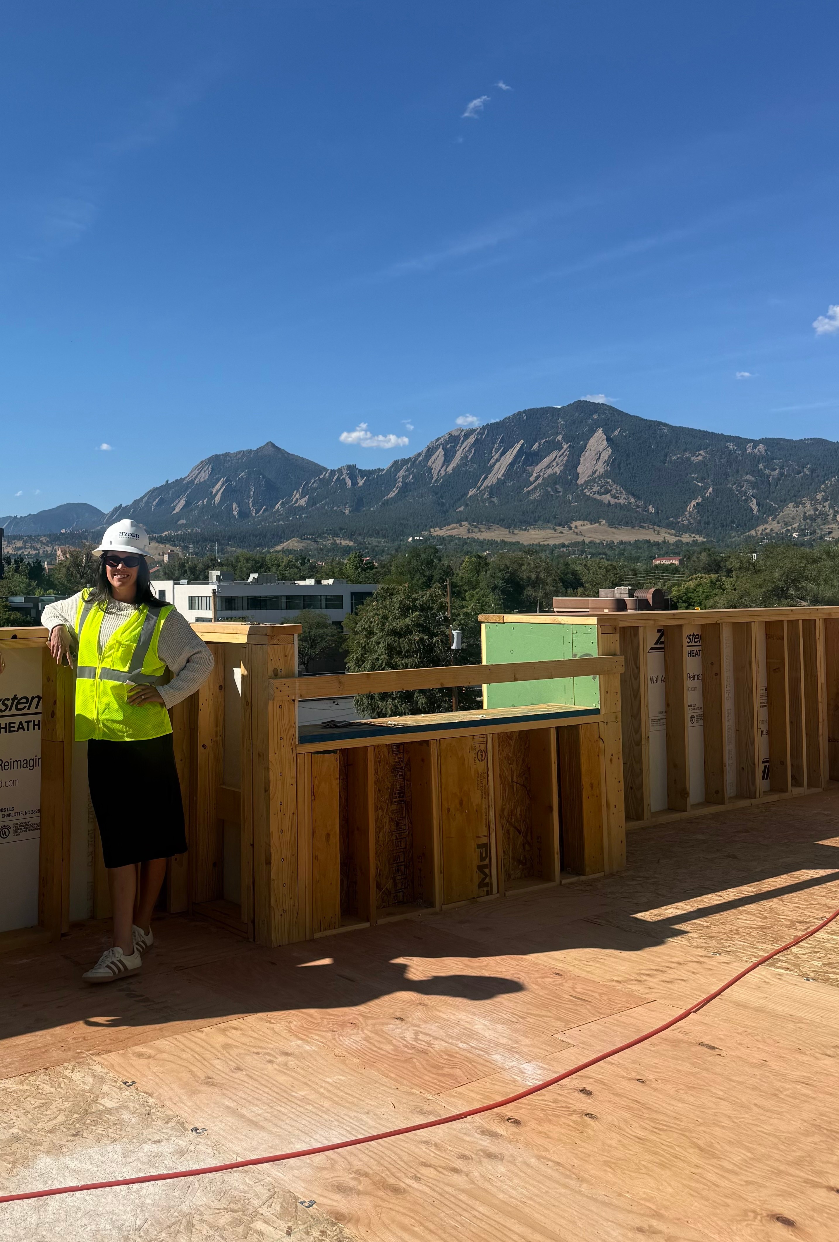 Person wearing a yellow safety vest and white hat standing on a wooden platform with mountains and blue sky in background.