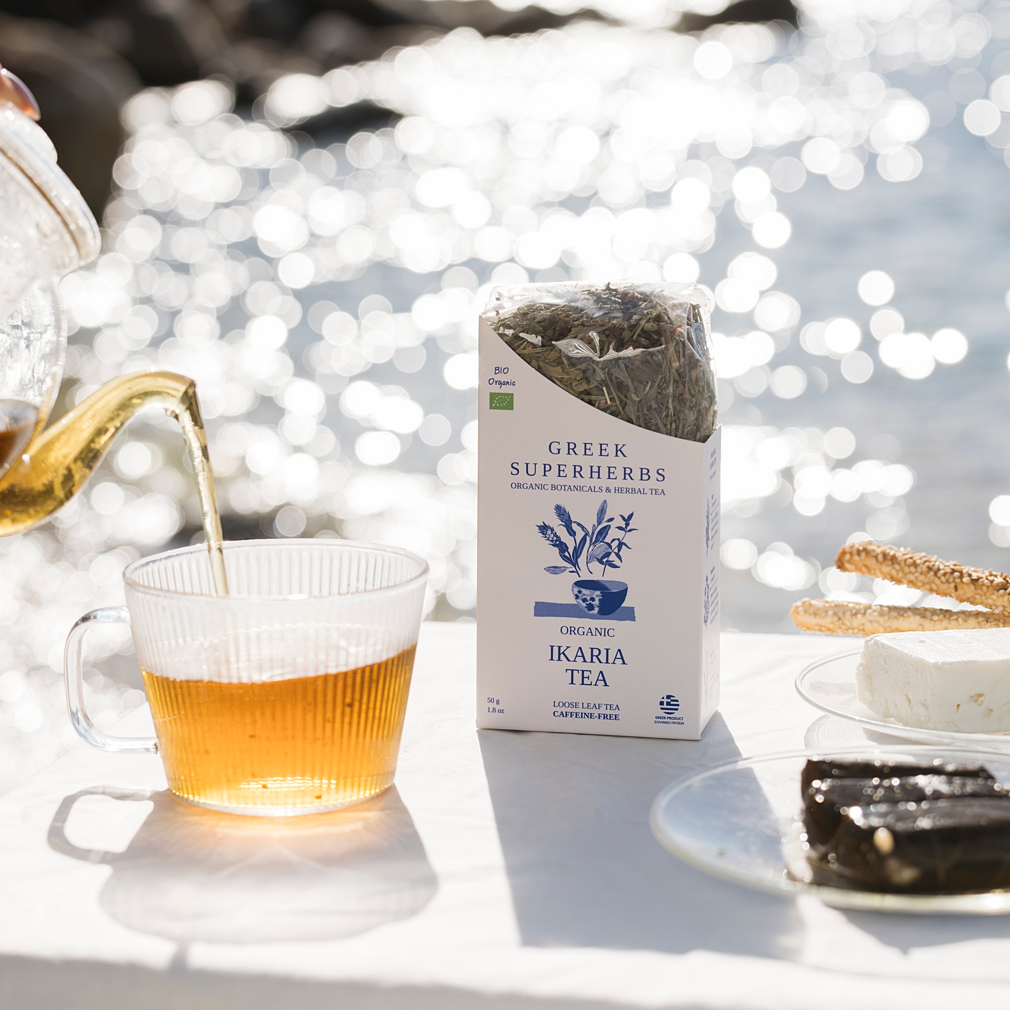 A glass of tea, a box of Greek superherbs, and snacks on a white surface near water with sunlight reflections.
