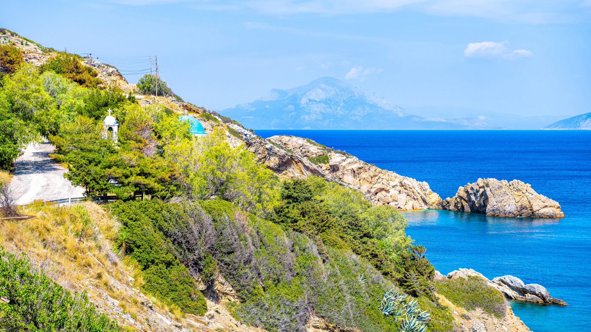 Coastal hillside with greenery, rocks, and blue sea in the background under a partly cloudy sky.