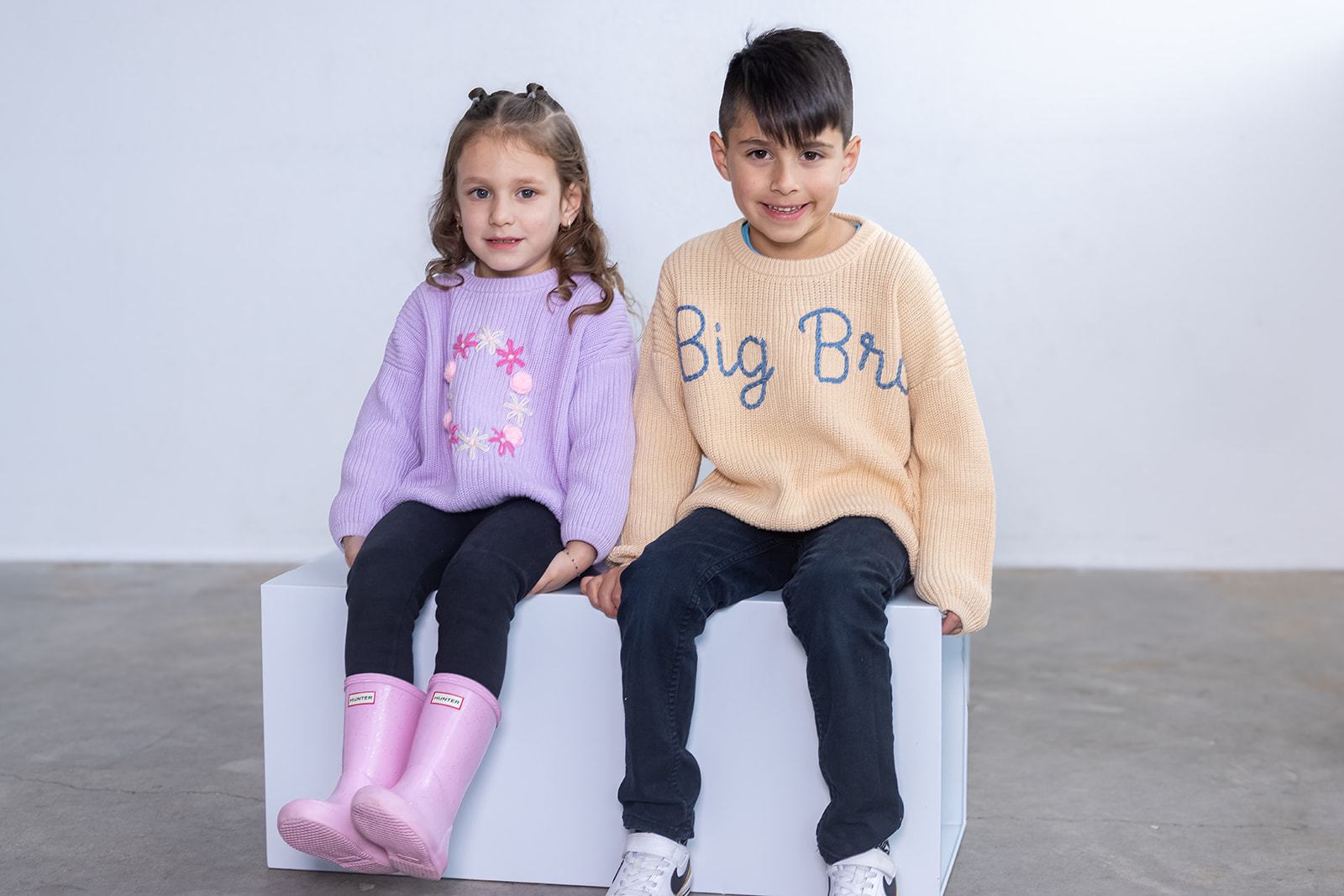 Two children sitting on a white block, girl in pink rain boots and purple sweater, boy in beige sweater and dark pants, smiling.