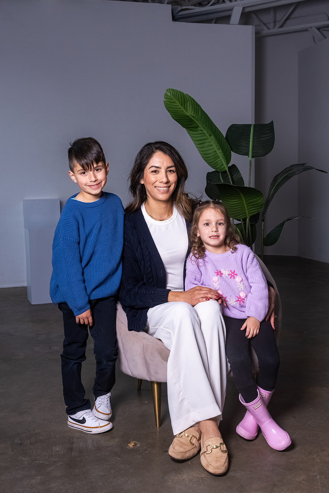 Woman sitting on a chair with two children, one standing and one sitting, in a room with a large plant.