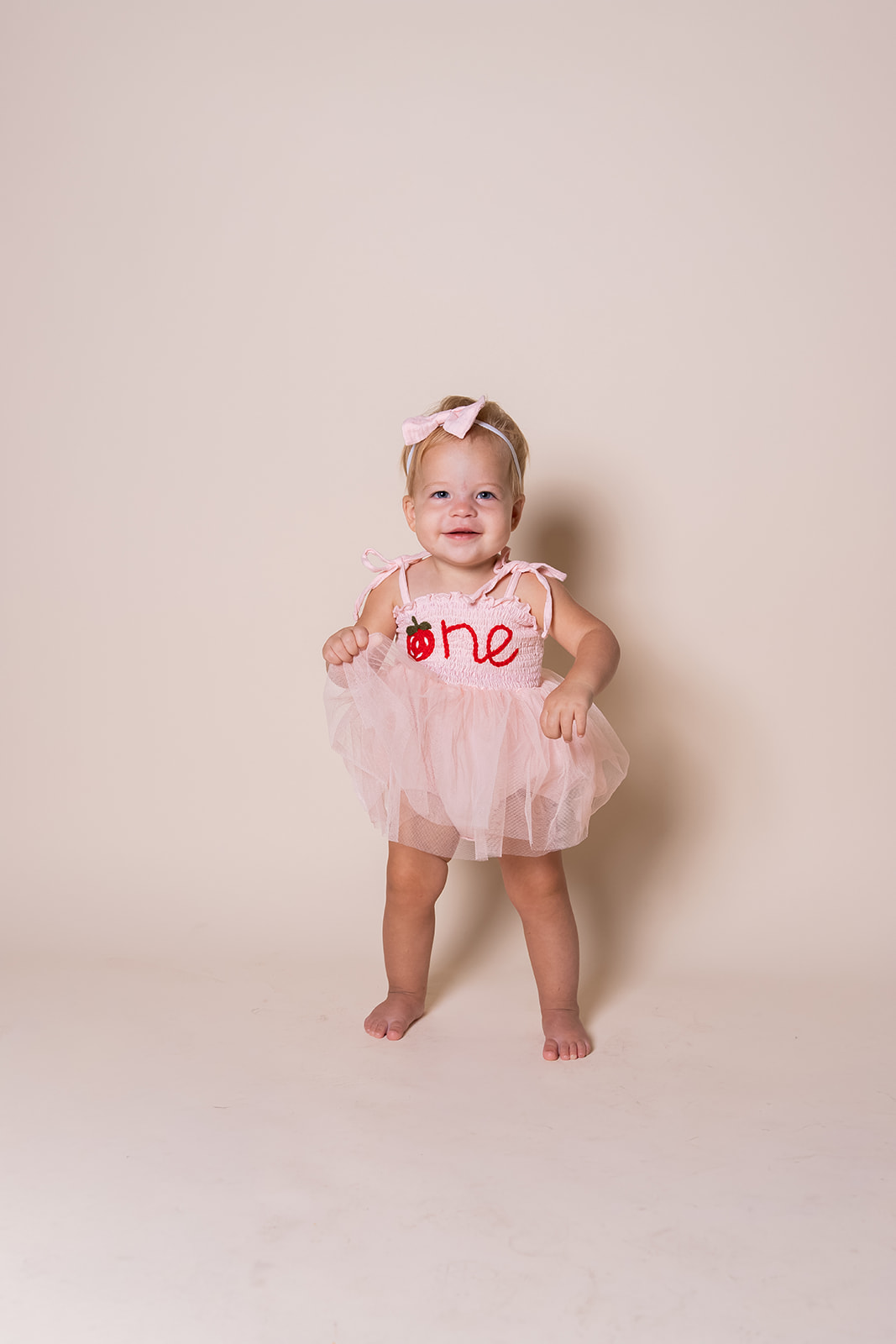 Young girl in a pink tutu dress with a bow in her hair, standing barefoot against a plain background.